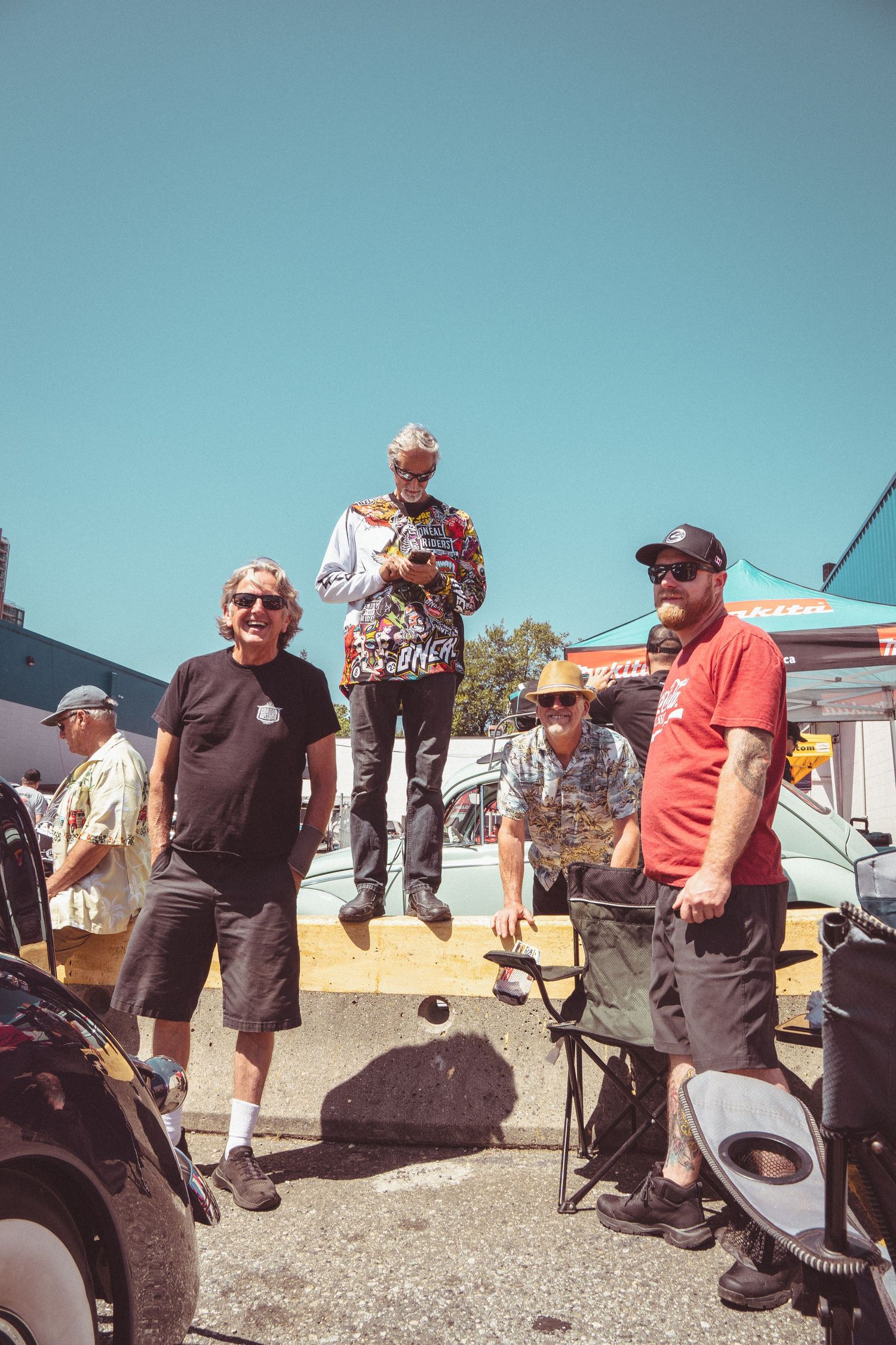 Group of four men standing outdoors at a sunny car show event with clear blue sky.