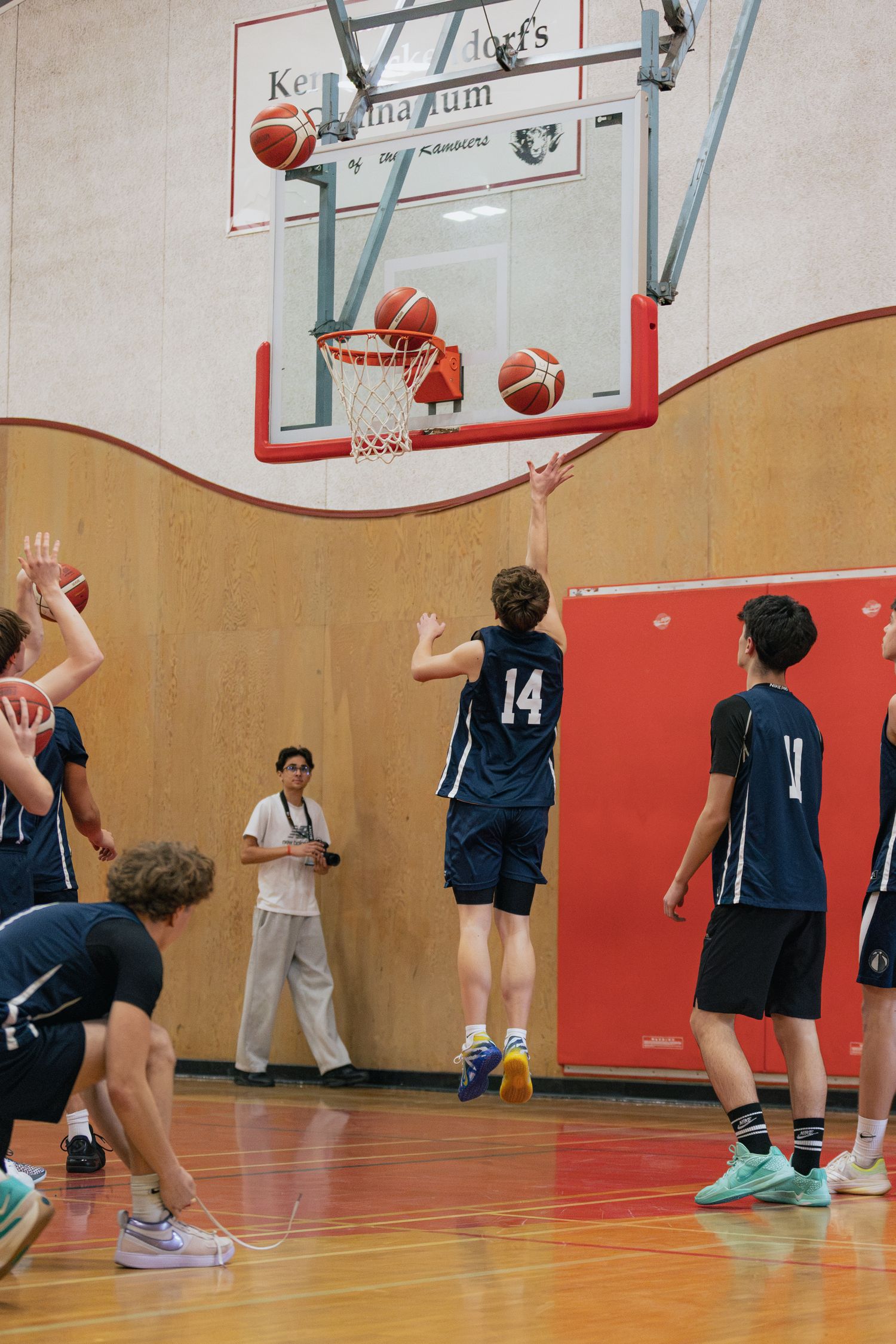 Youth basketball players in navy uniforms practice shooting drills in a school gymnasium.
