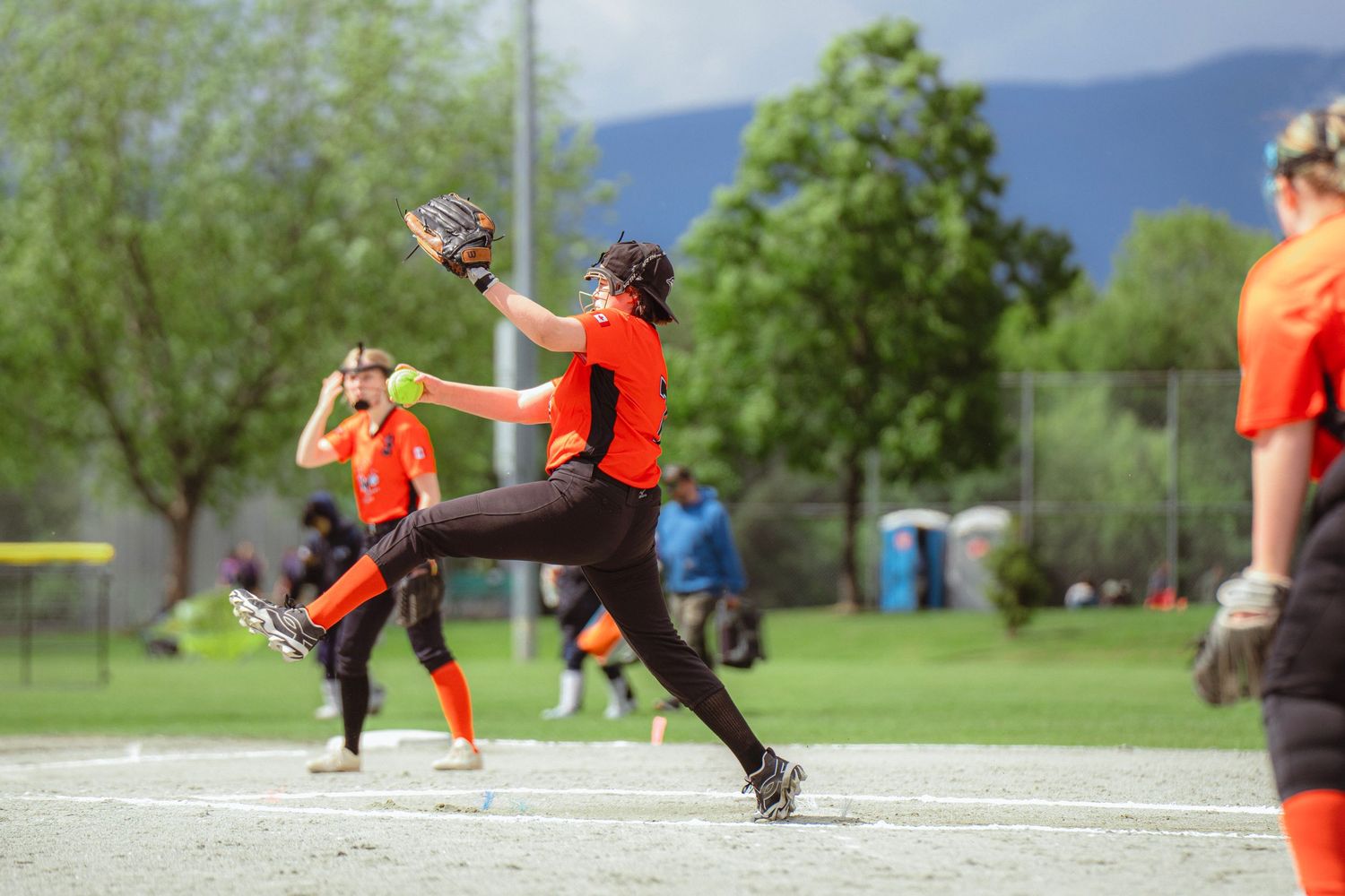 Female softball pitcher in orange uniform mid-windup on outdoor field with mountains in background.