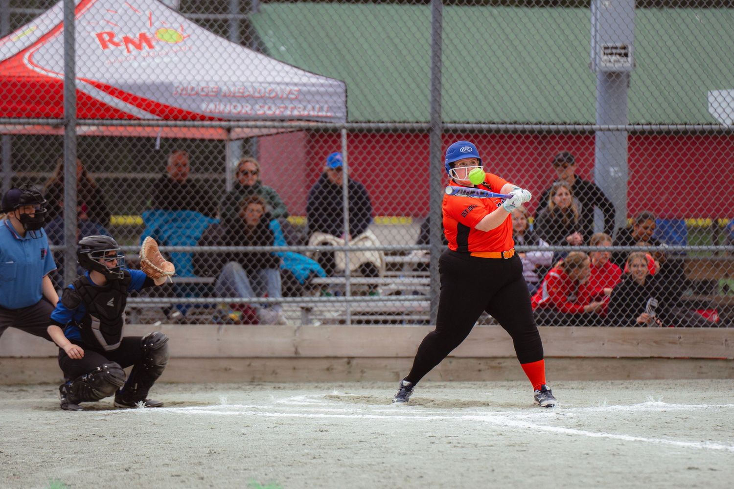 Female softball batter in orange uniform at plate with catcher and umpire behind her.