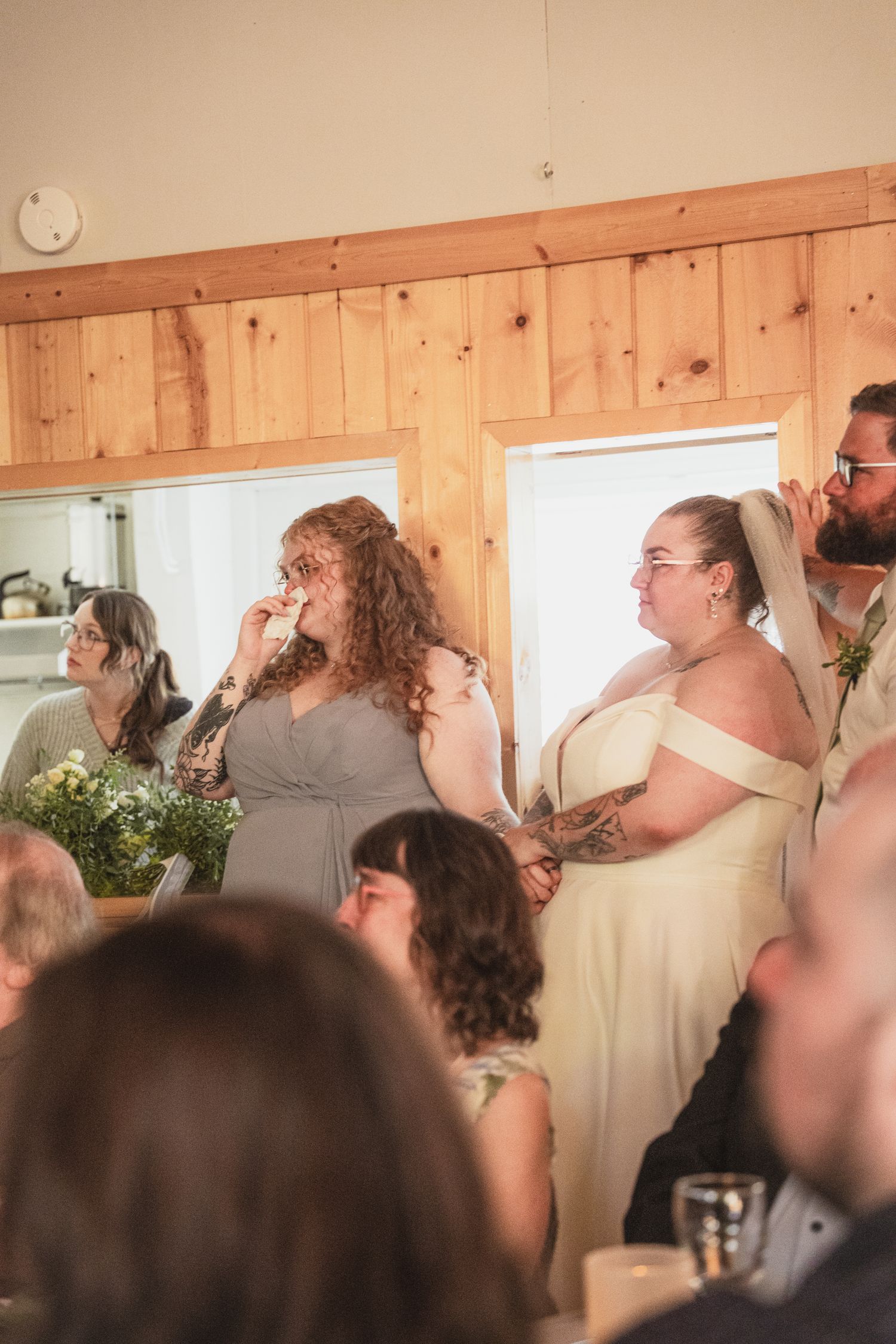 Emotional wedding guests including a bridesmaid wiping tears during a heartfelt reception speech at a rustic venue.