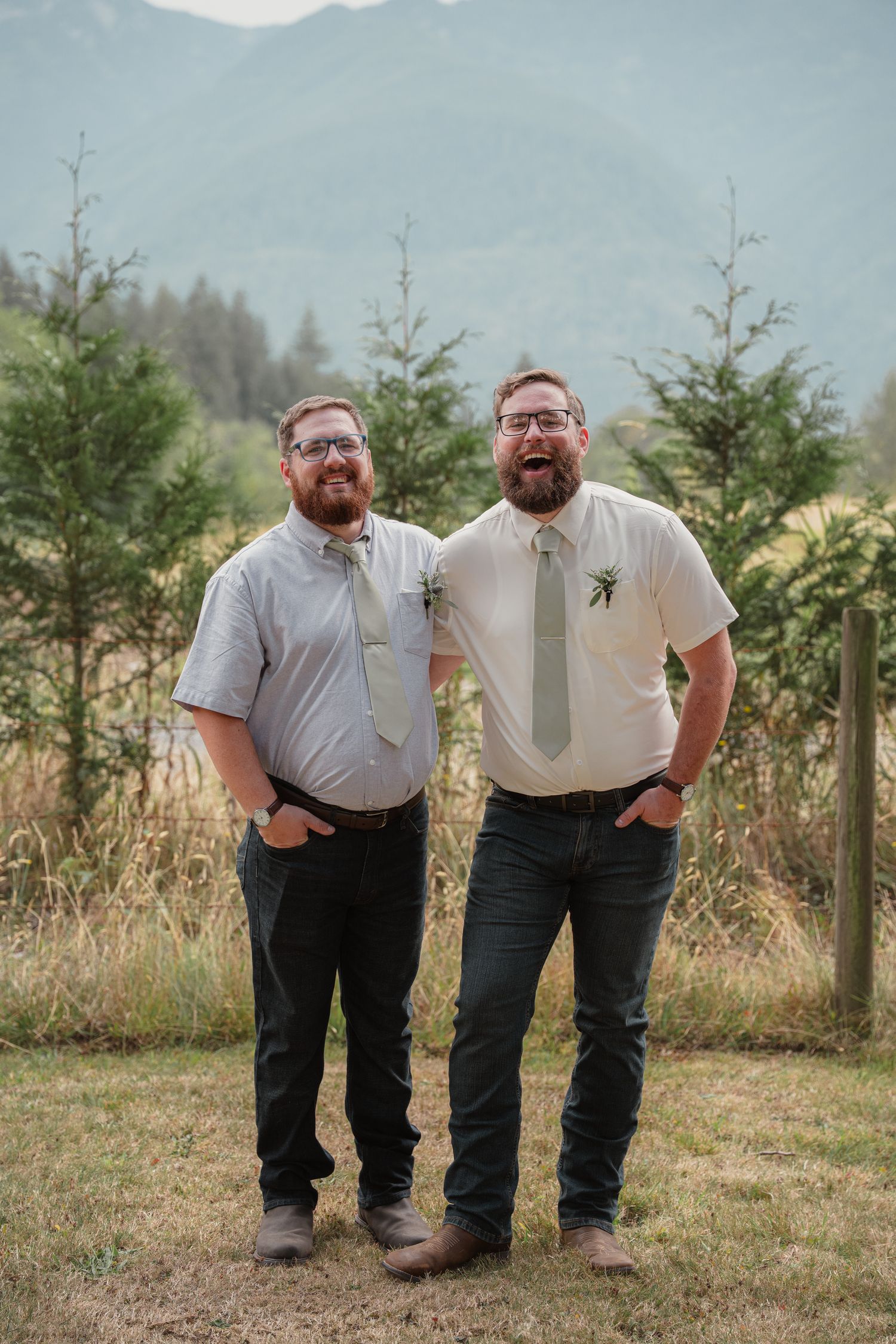 Two bearded grooms in ties and jeans laugh together outdoors with mountains and evergreen trees behind them.