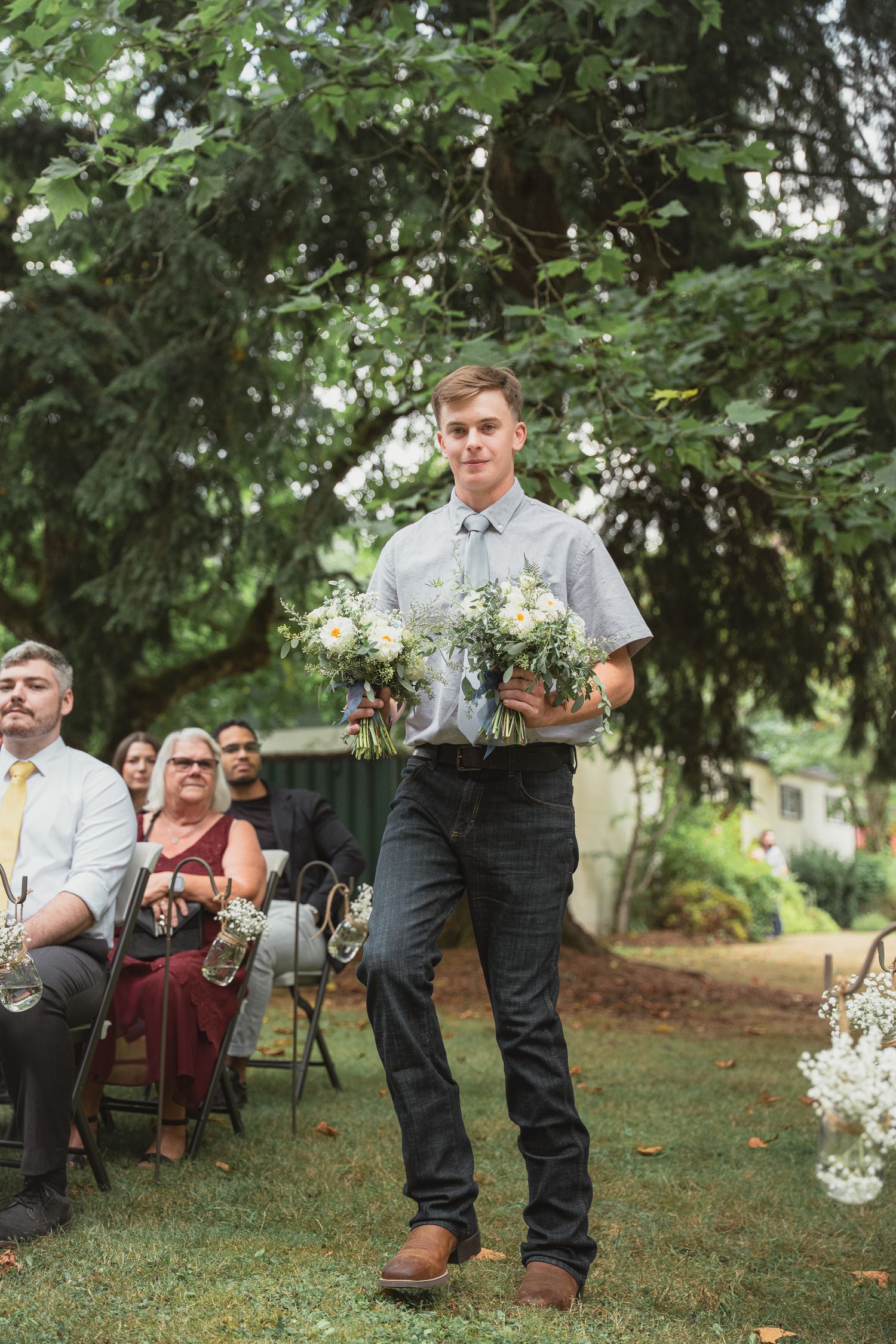 Young groomsman carrying white floral bouquets down outdoor wedding aisle, wearing gray shirt and dark jeans.