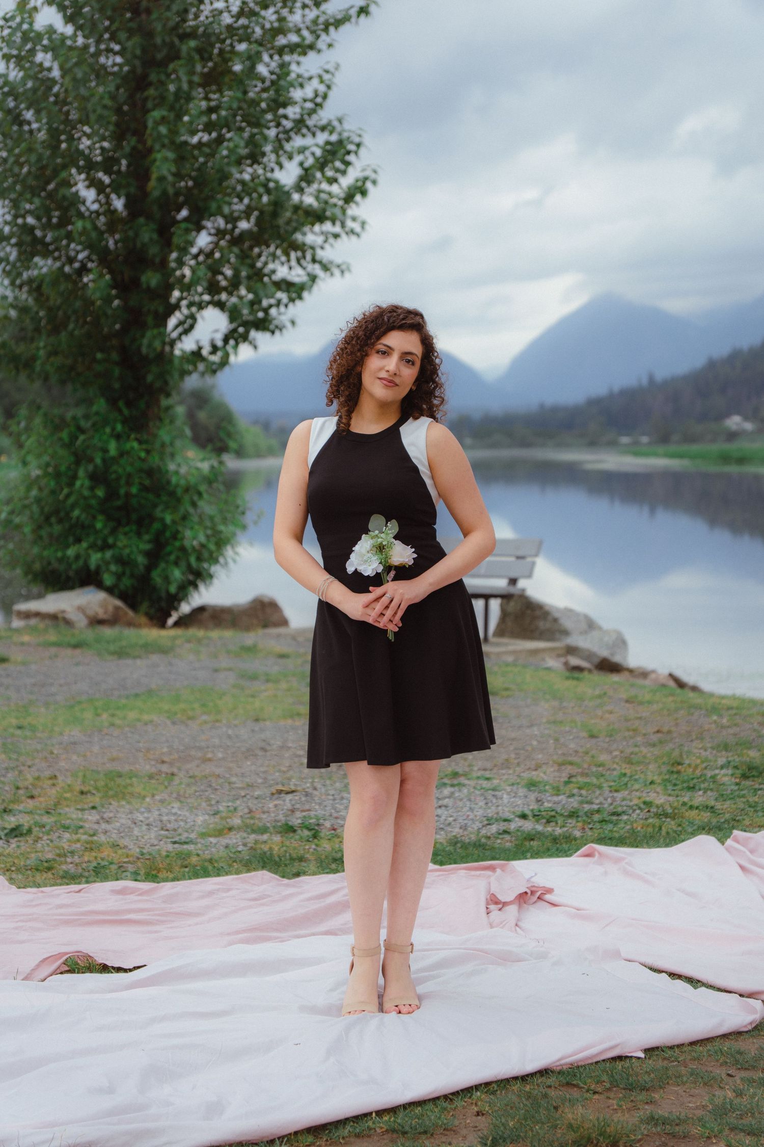 Woman in black dress holding white bouquet standing on pink fabric by a reflective mountain lake.