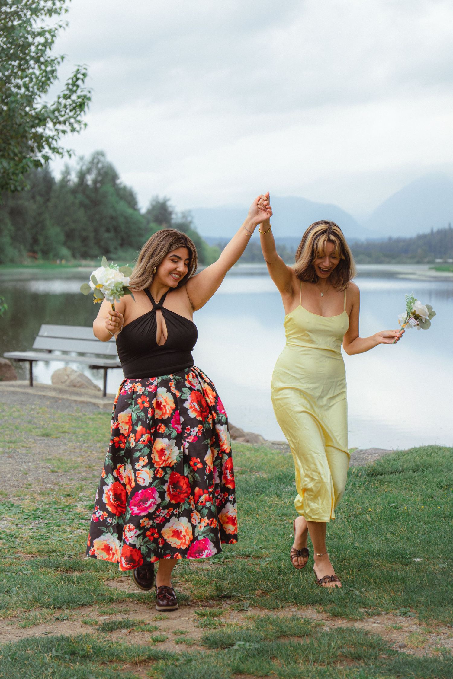 Two women in floral and yellow dresses dance and hold hands by a lakeside.