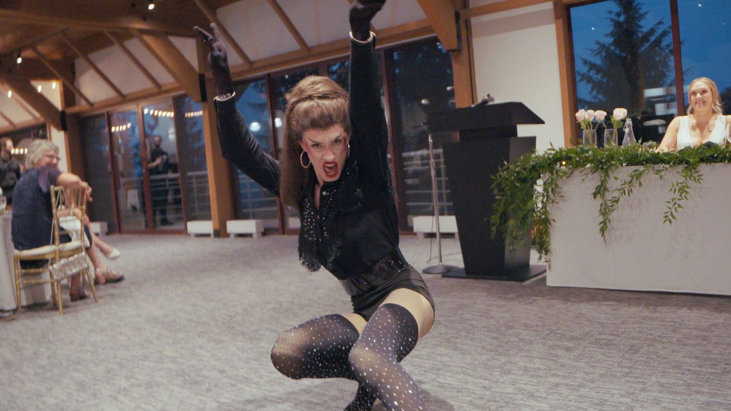 A performer in a black sequined outfit does a dramatic split pose at an elegant indoor wedding reception.