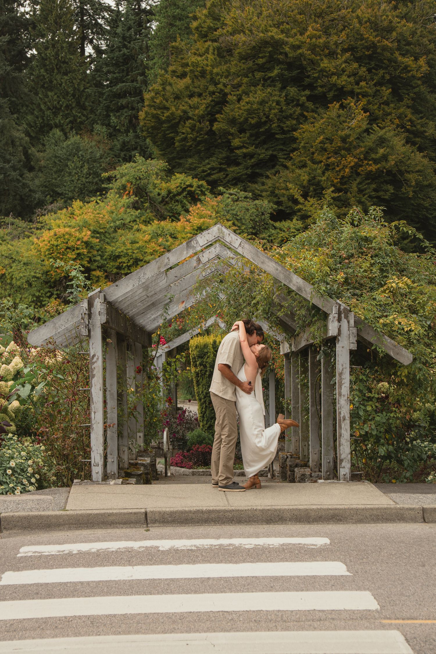 Couple sharing a romantic kiss under a rustic wooden arbor surrounded by lush garden greenery on their wedding day.