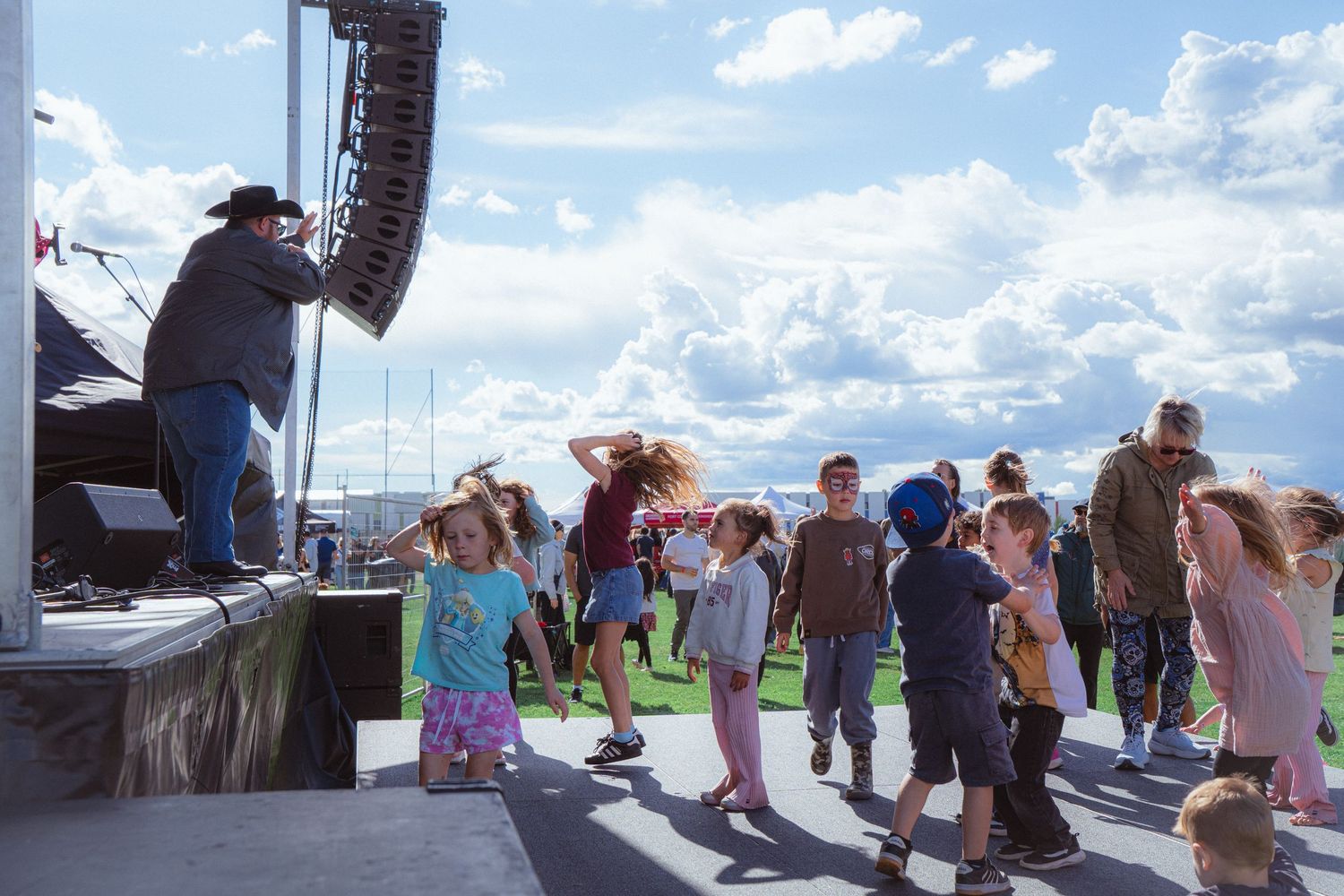 Children dancing in front of outdoor festival stage under dramatic cloudy sky on sunny day.