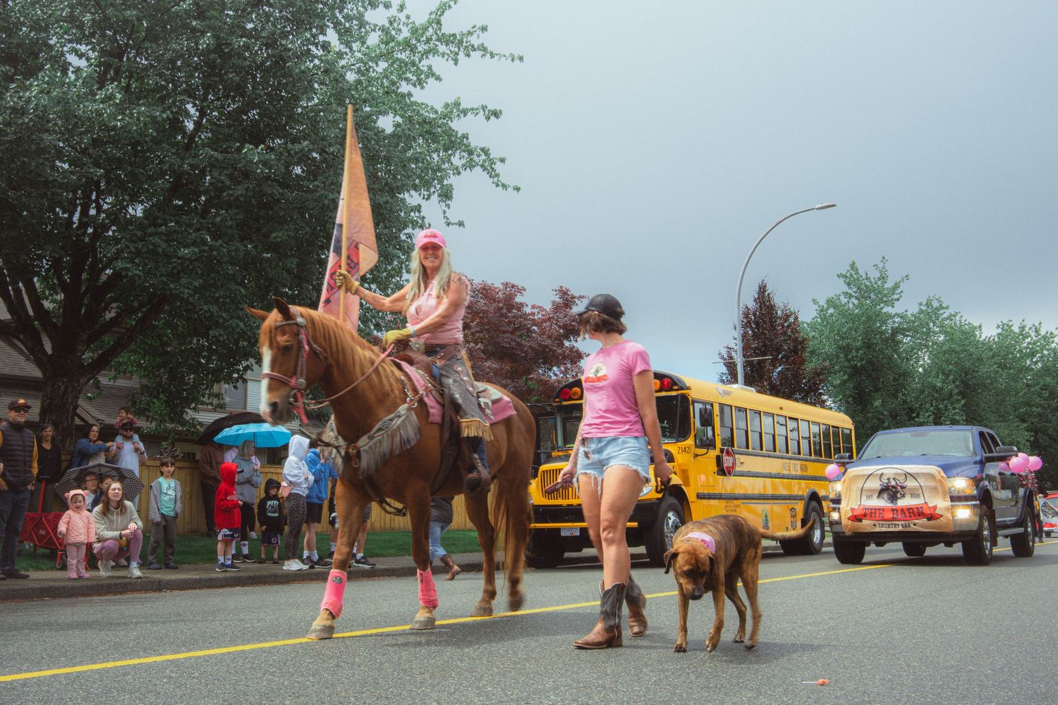 A woman on horseback with a flag leads a dog walker past a school bus in a rainy parade.