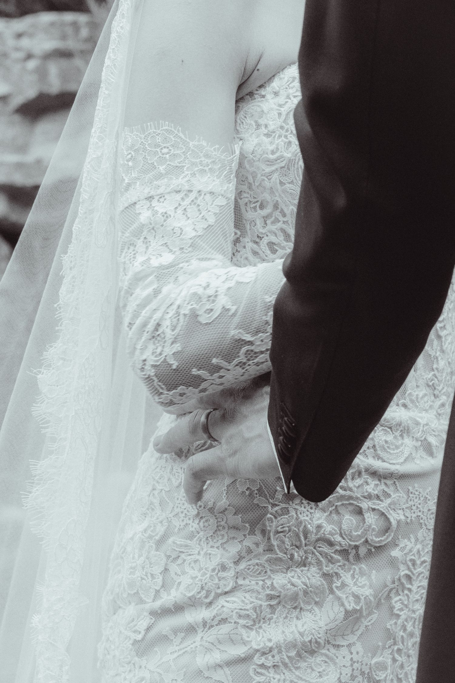 Black and white photo of bride and groom holding hands, showcasing intricate lace wedding dress and veil details.