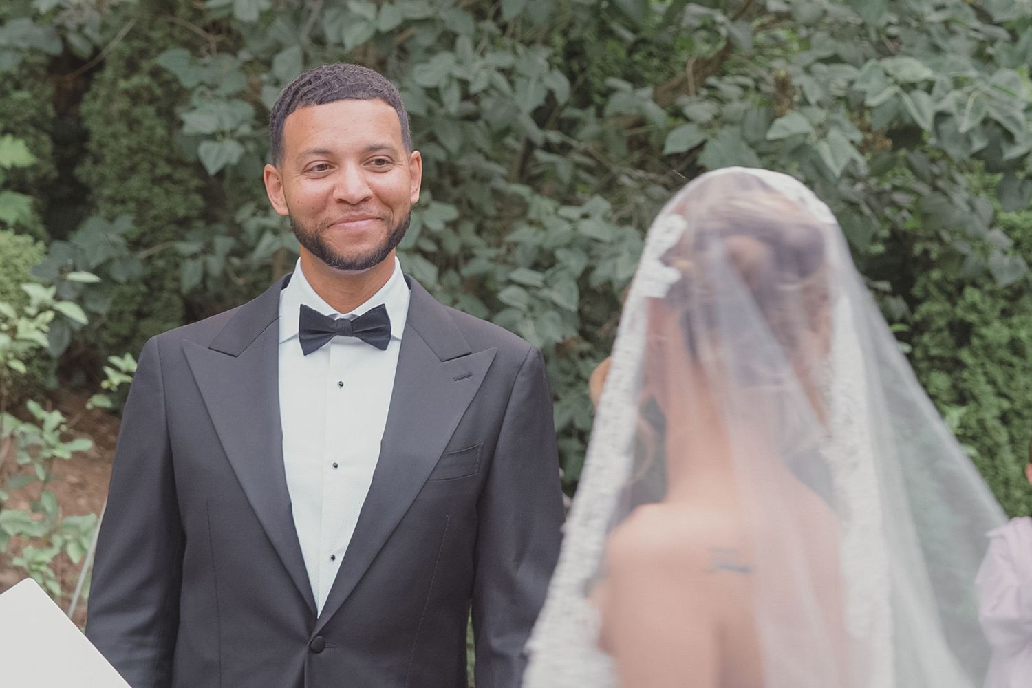 Groom in black tuxedo smiling at bride in wedding veil during outdoor ceremony.