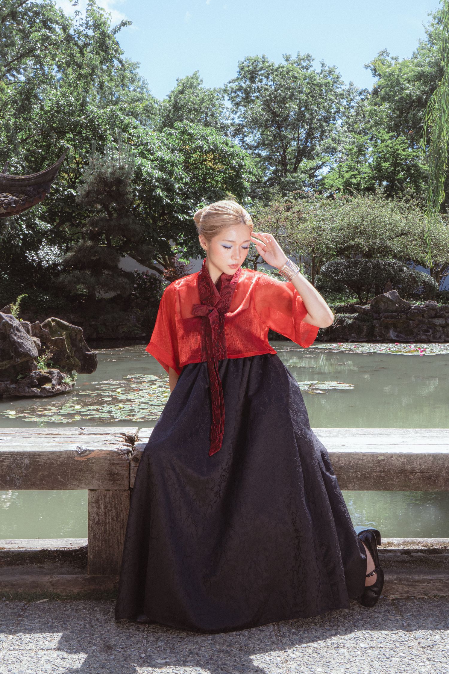 Woman in red top and black skirt poses gracefully in a serene garden with a pond and lush greenery.