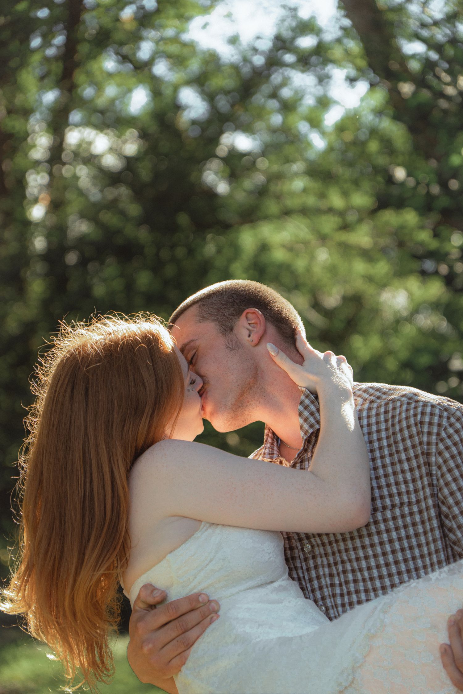 A romantic couple shares a tender kiss outdoors, the woman with long red hair embracing a man in a plaid shirt.