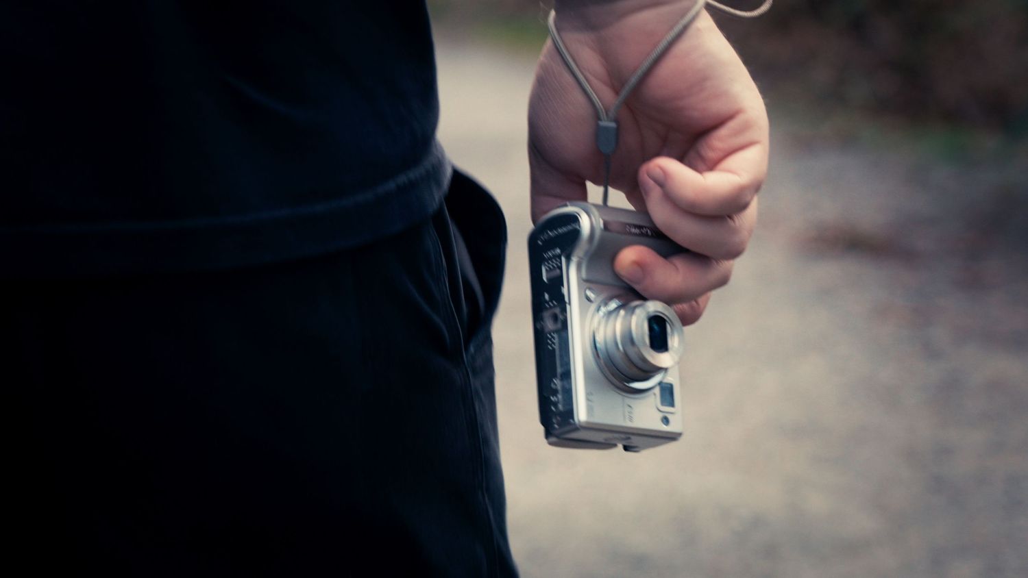 Hand holding a compact silver digital camera with a strap against a dark clothing background outdoors.