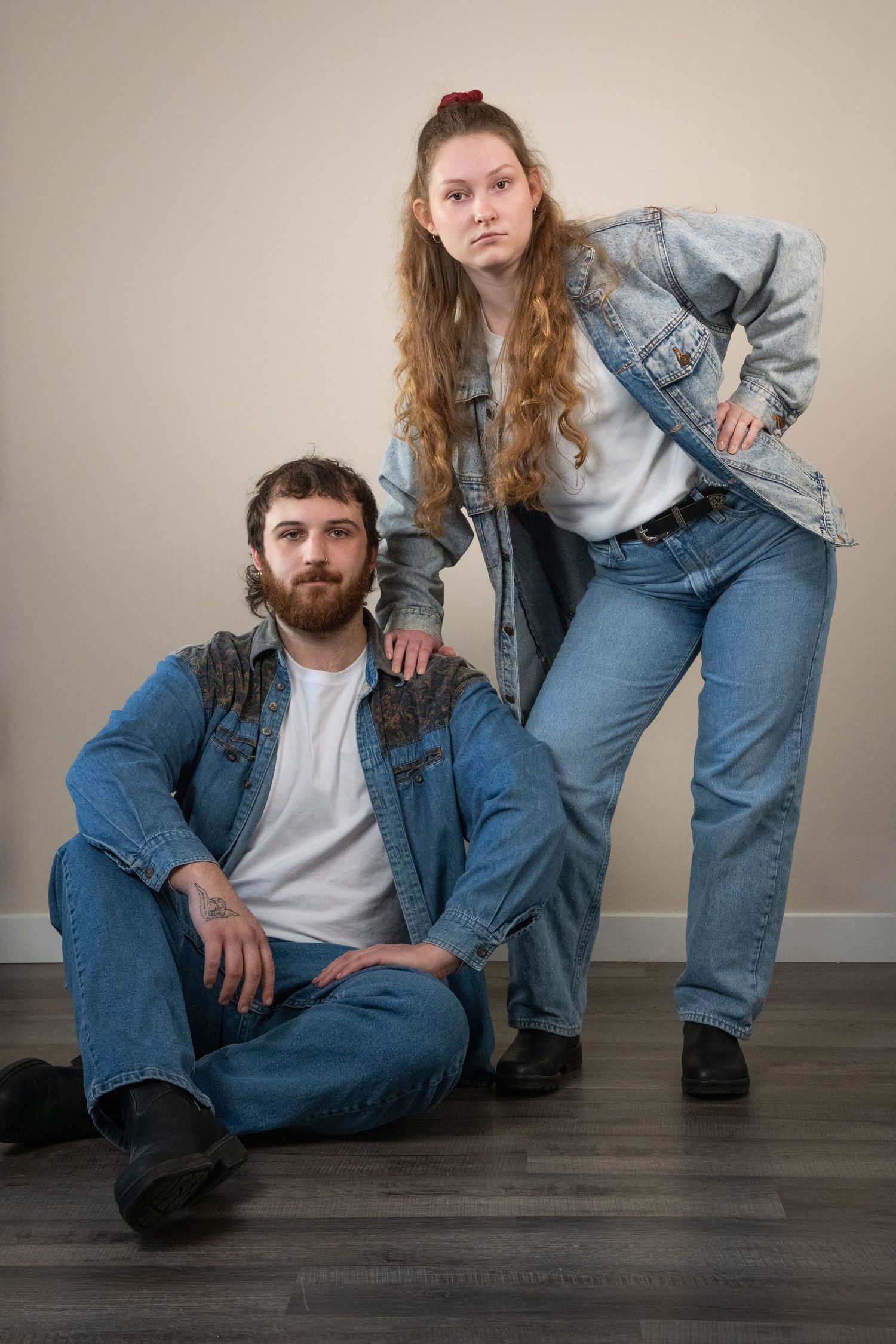 A bearded man sits on the floor while a woman in denim jacket stands behind him, both wearing all-denim outfits.