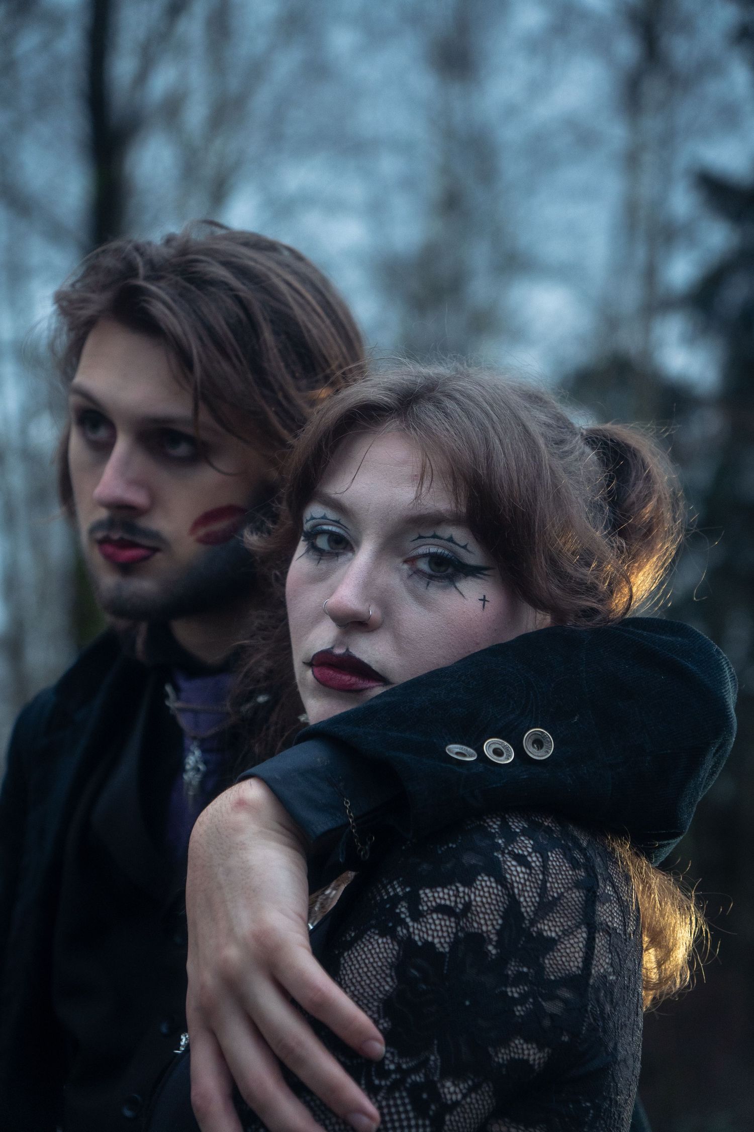 A gothic couple in dark theatrical makeup embrace outdoors, the woman wearing black lace in a moody forest setting.