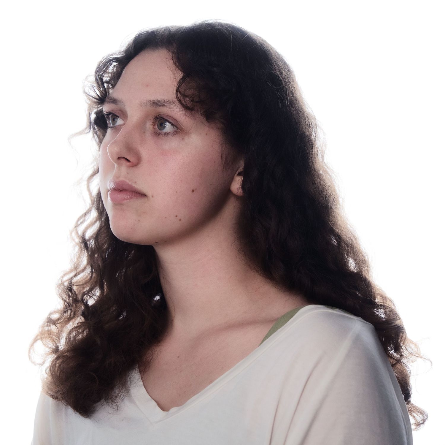 Young woman with long curly dark hair in white v-neck top gazing upward against white background.