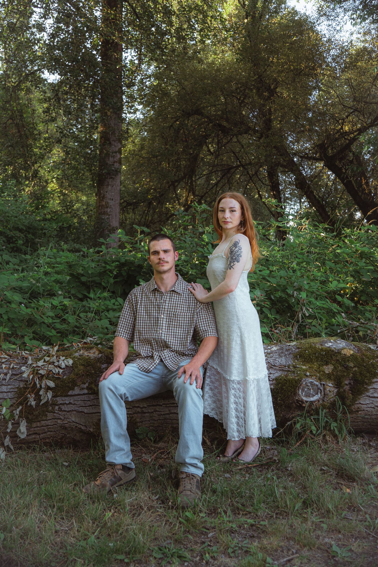 Couple posing in a forest, man sitting on mossy log, woman in white lace dress standing beside him.