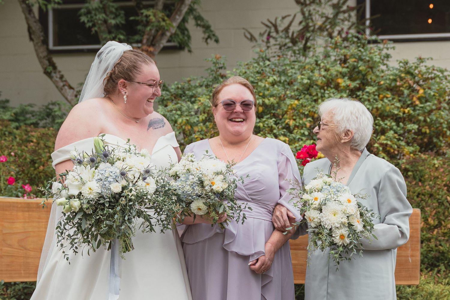 Bride in white gown with bridesmaids holding white floral bouquets smiling outdoors at a wedding ceremony.