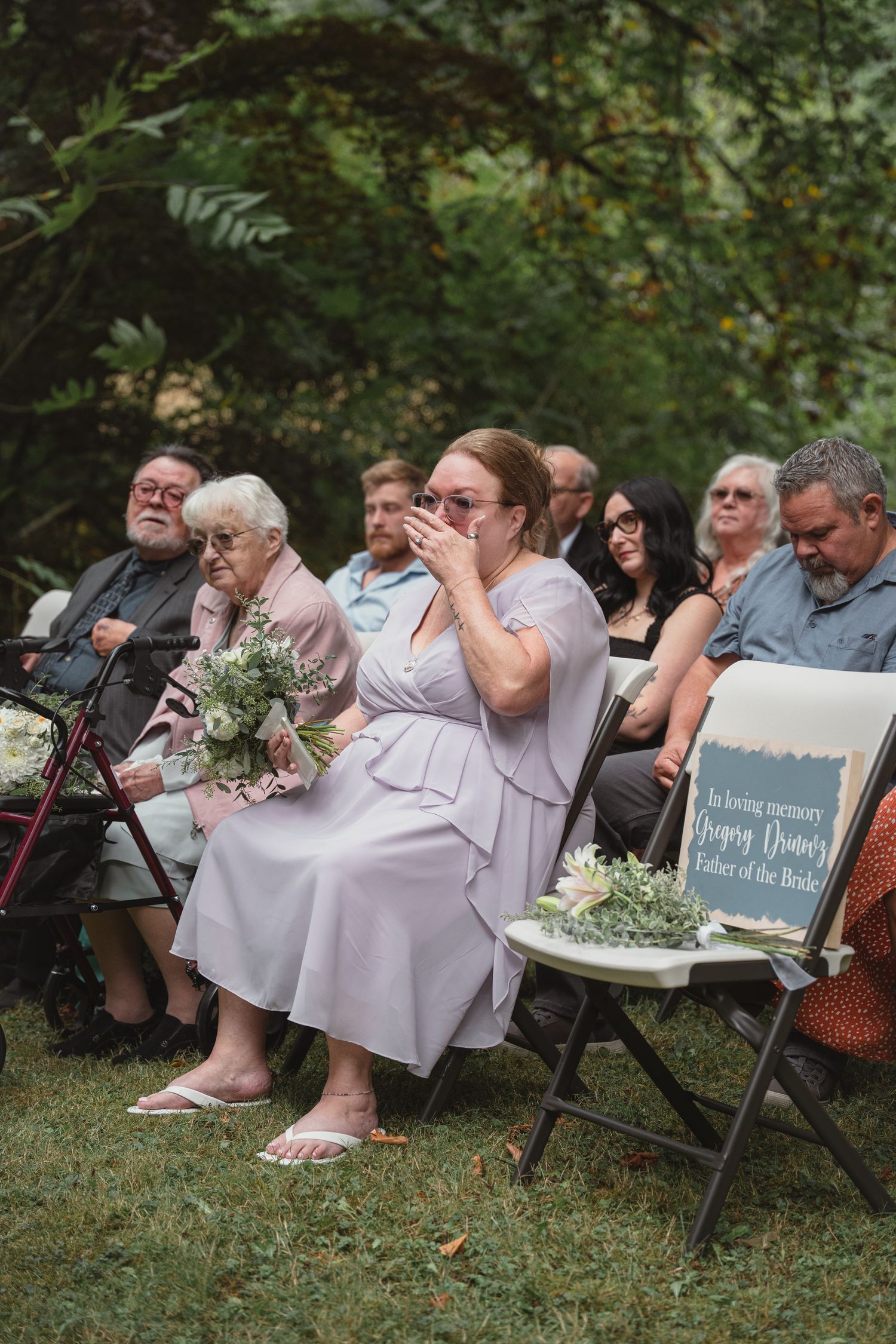 Emotional wedding guest in lavender dress wipes tears while seated at outdoor garden wedding ceremony.