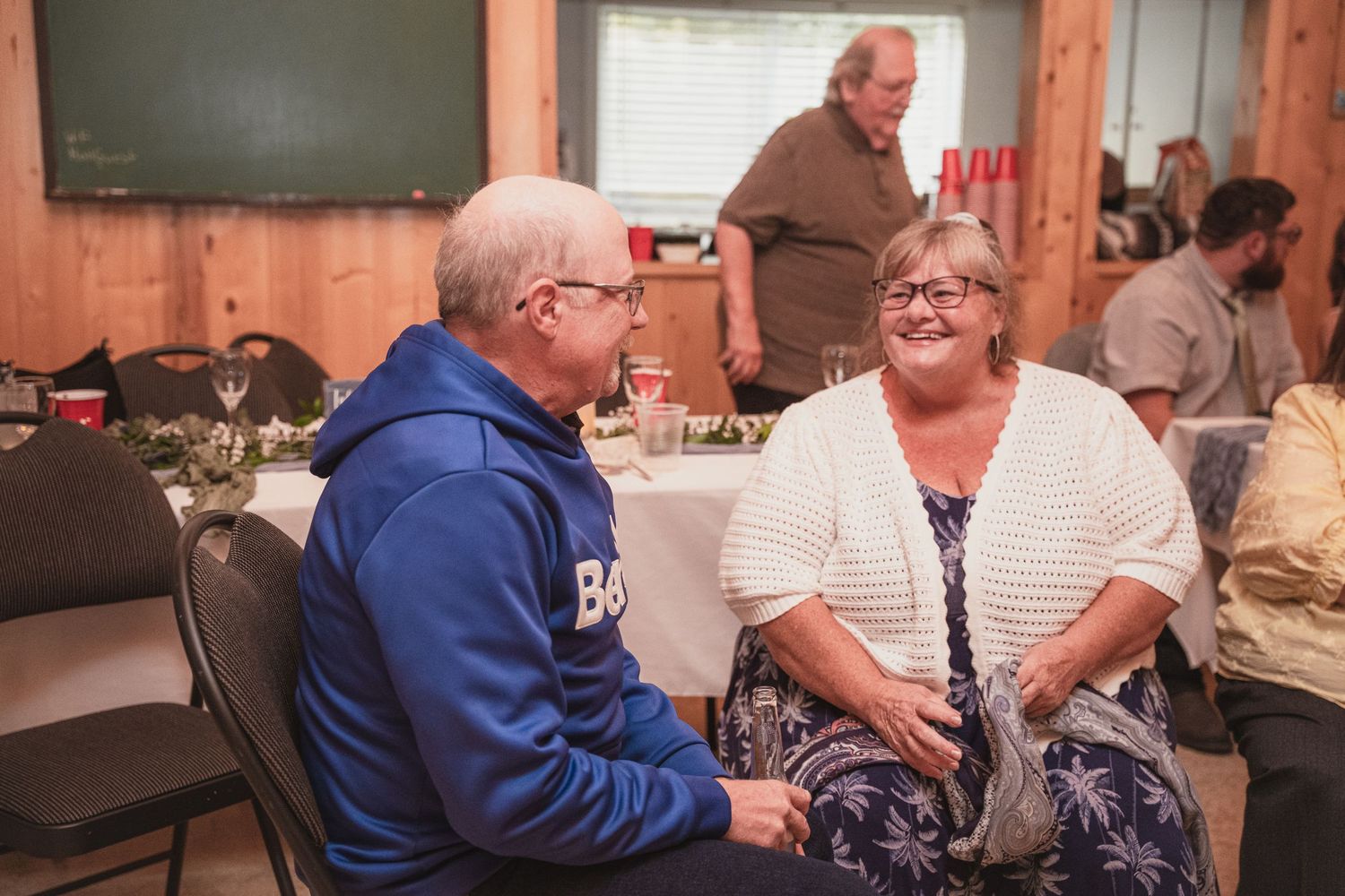 Two older wedding guests share a warm conversation while seated at a rustic barn wedding reception.