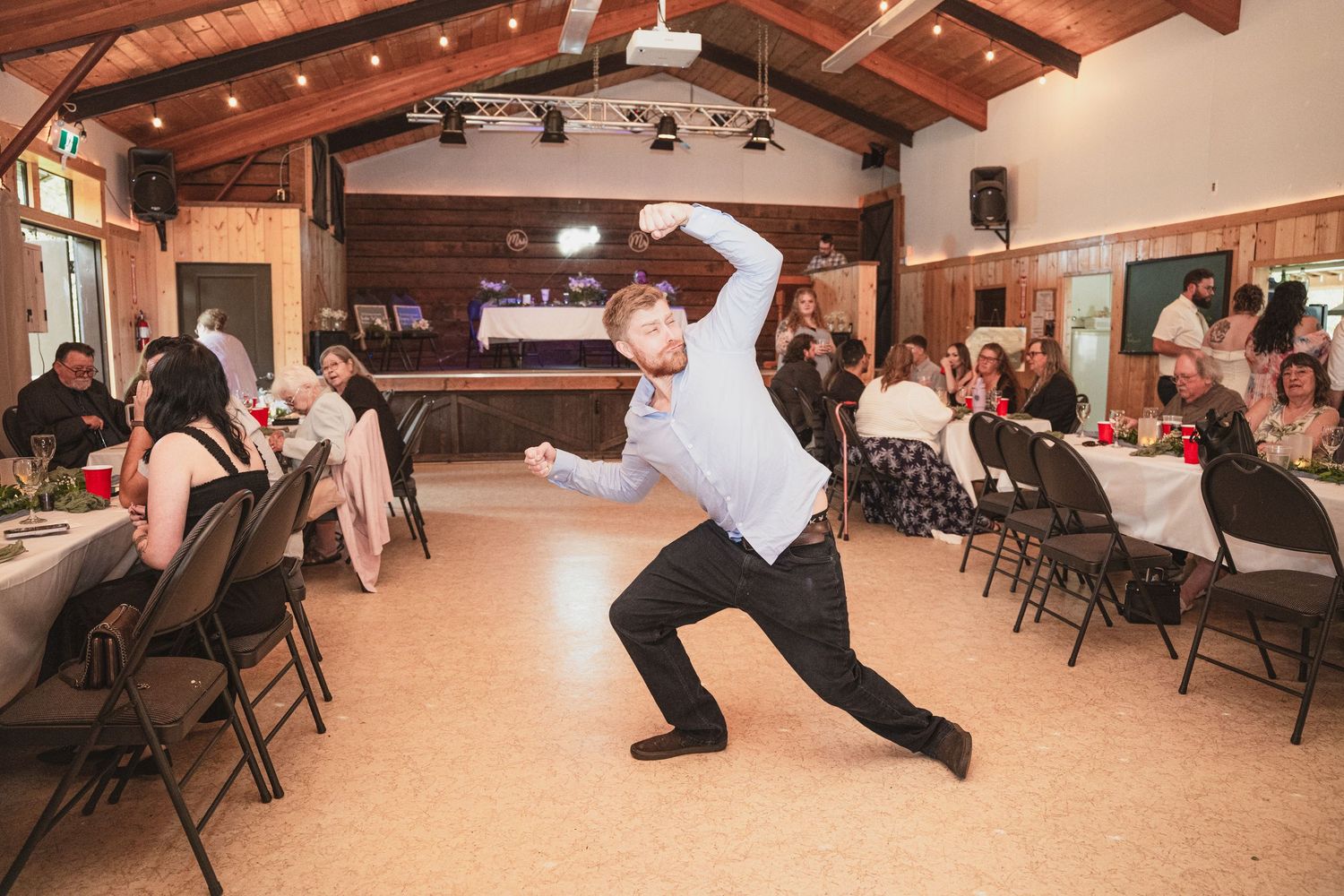 Man performing dramatic dance move at wedding reception in rustic hall with guests seated at tables.