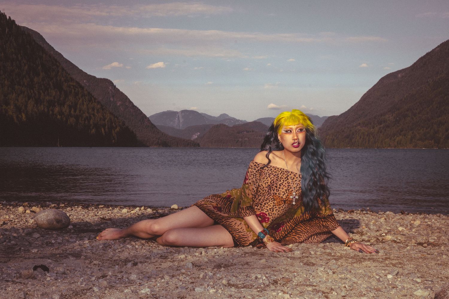 Woman with yellow hair and long dark braids poses on rocky lakeshore with mountain backdrop at dusk.