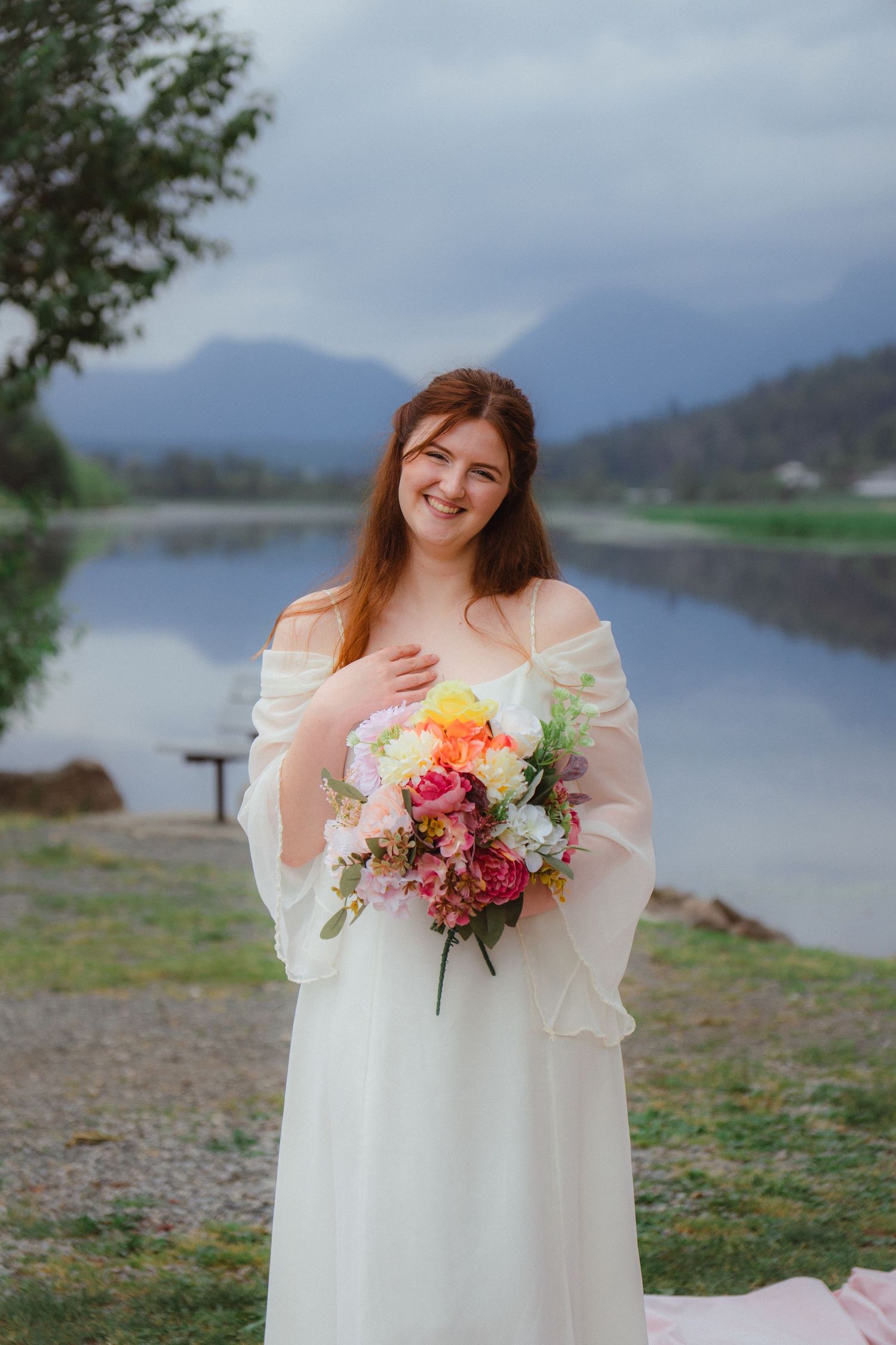 Smiling bride in white dress holds vibrant floral bouquet near a misty mountain lake.