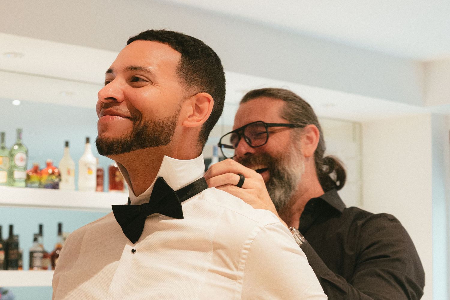 Man in white tuxedo shirt smiles as another man adjusts his black bow tie indoors near a bar.