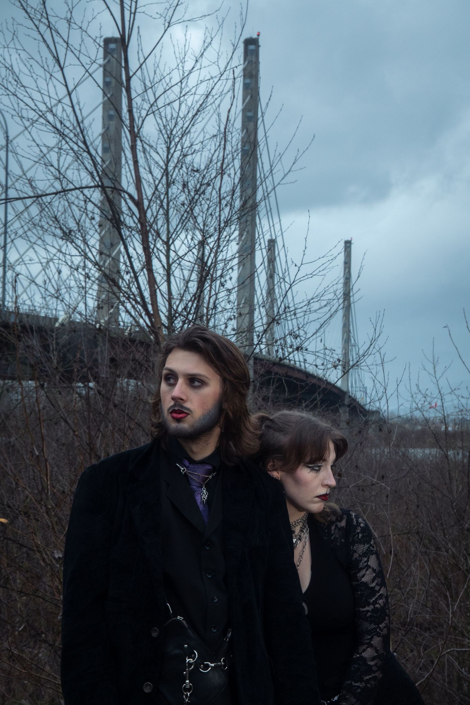 Gothic couple in black attire pose near bare trees and industrial structures under cloudy sky.