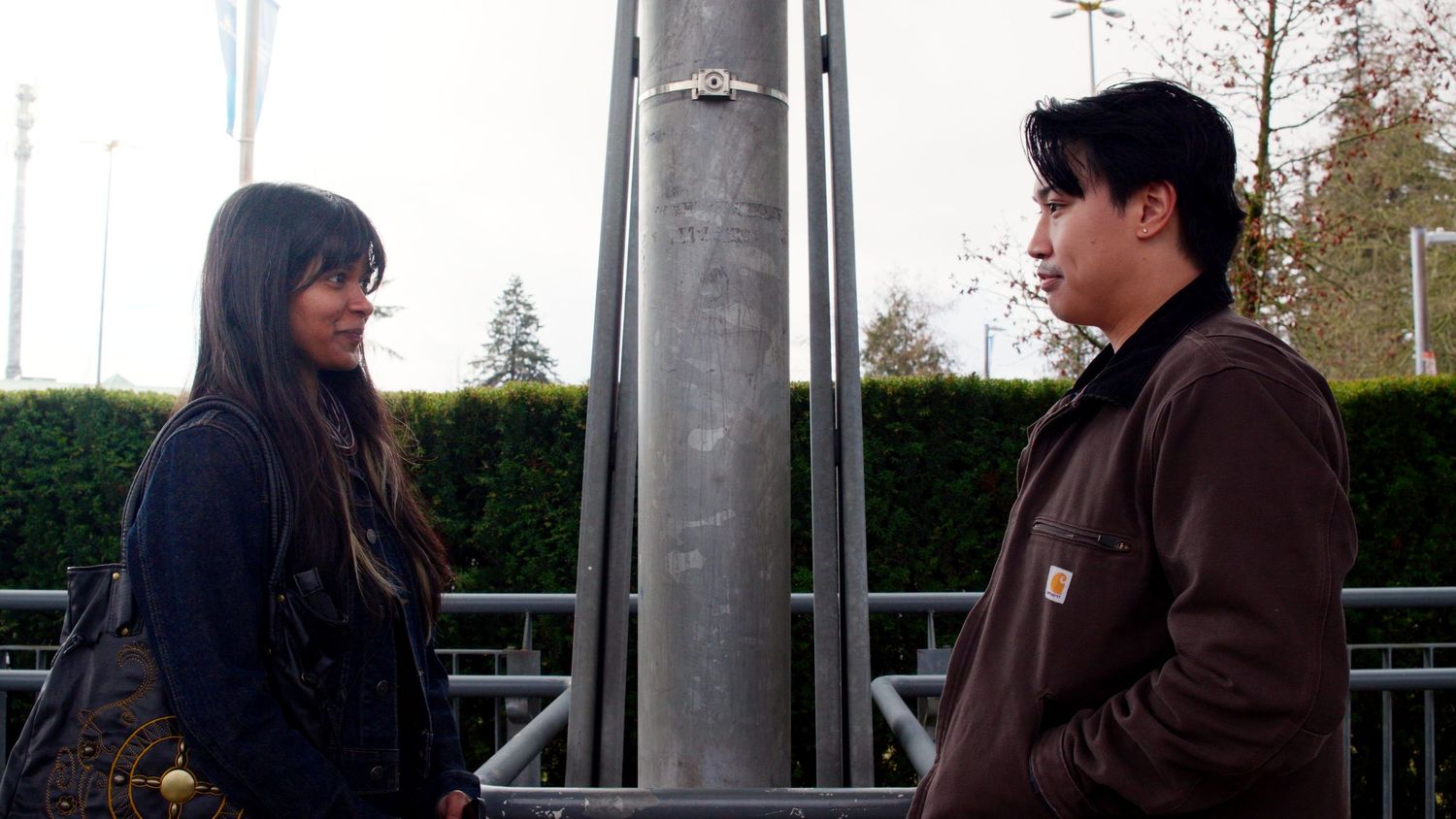 Two young people stand facing each other outdoors near a metal pole in a dramatic scene.