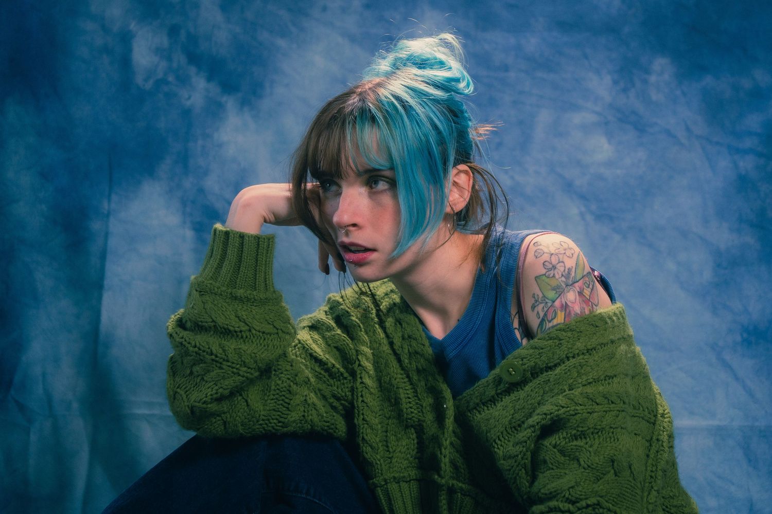 Young woman with blue hair and shoulder tattoo in green knit cardigan poses in moody blue studio.