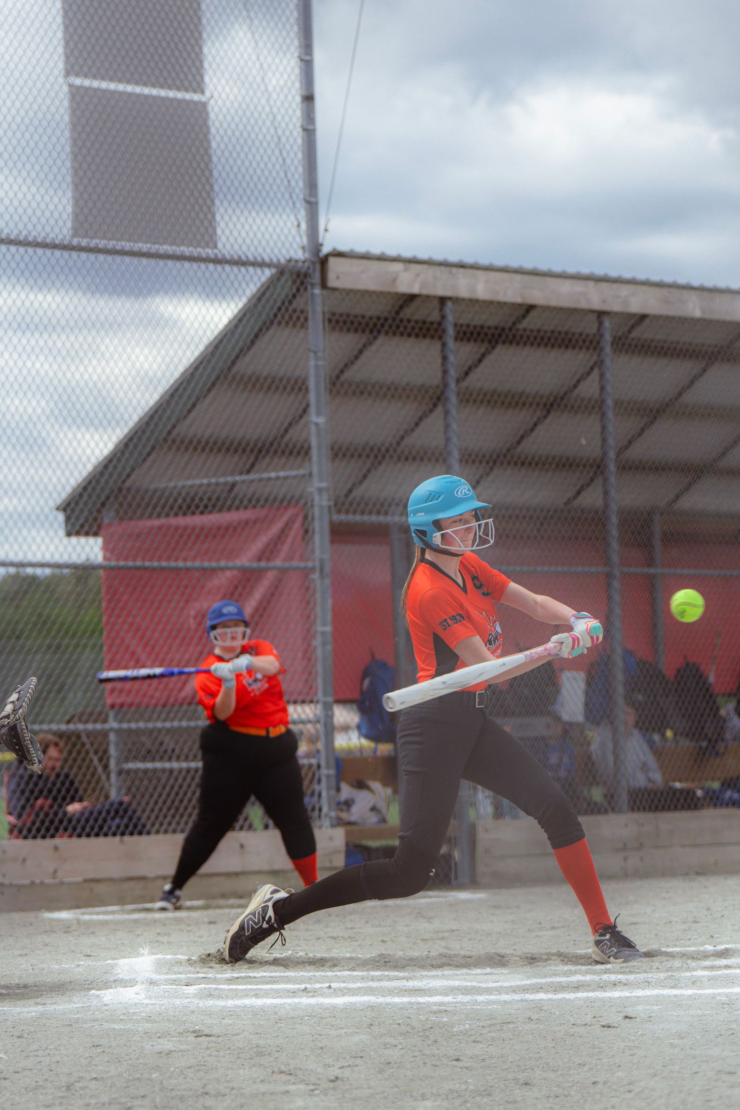 Female softball player in orange uniform swinging at a yellow ball during a game.