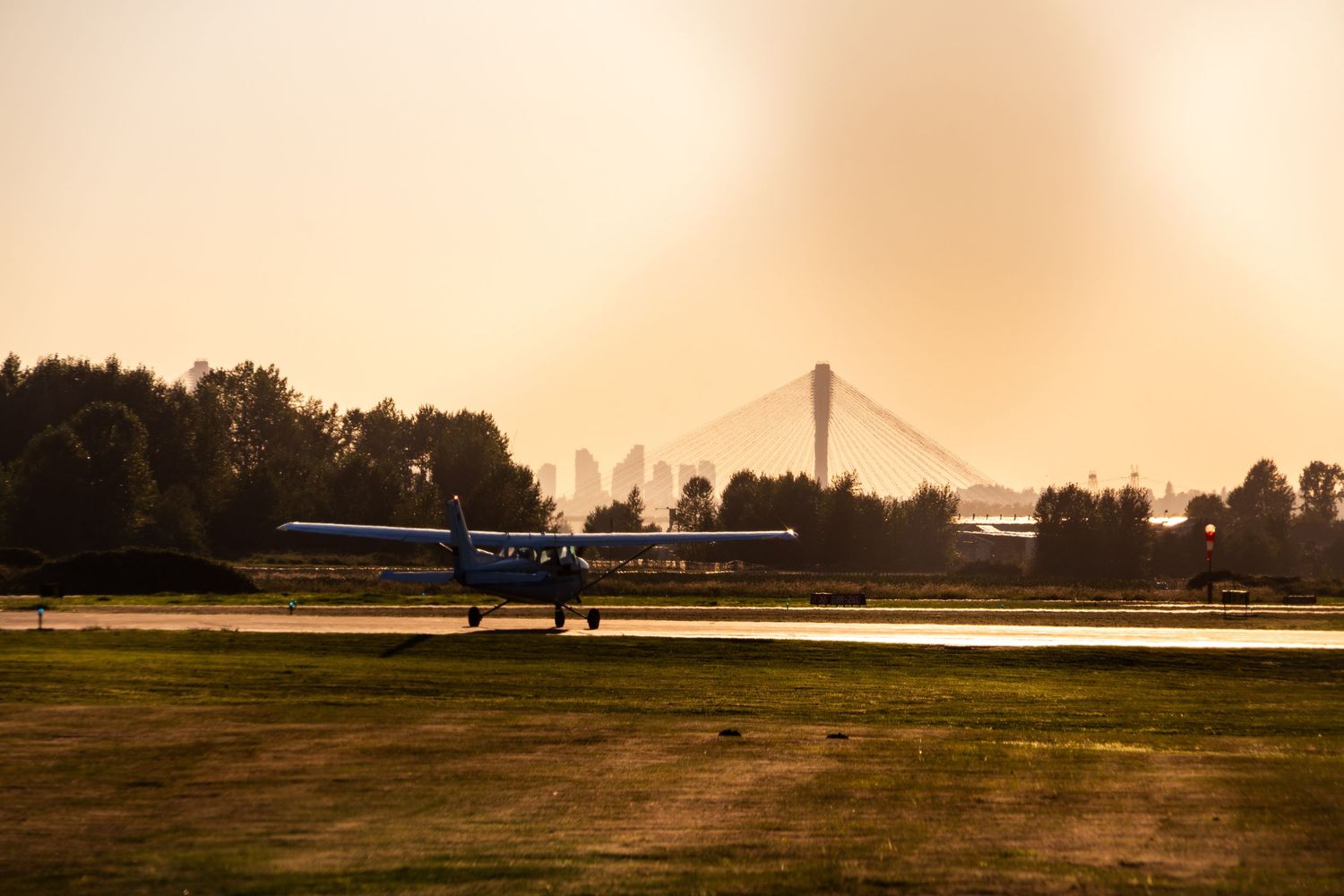 A small propeller plane taxis on a grassy airfield at sunset with a cable-stayed bridge visible in the background.