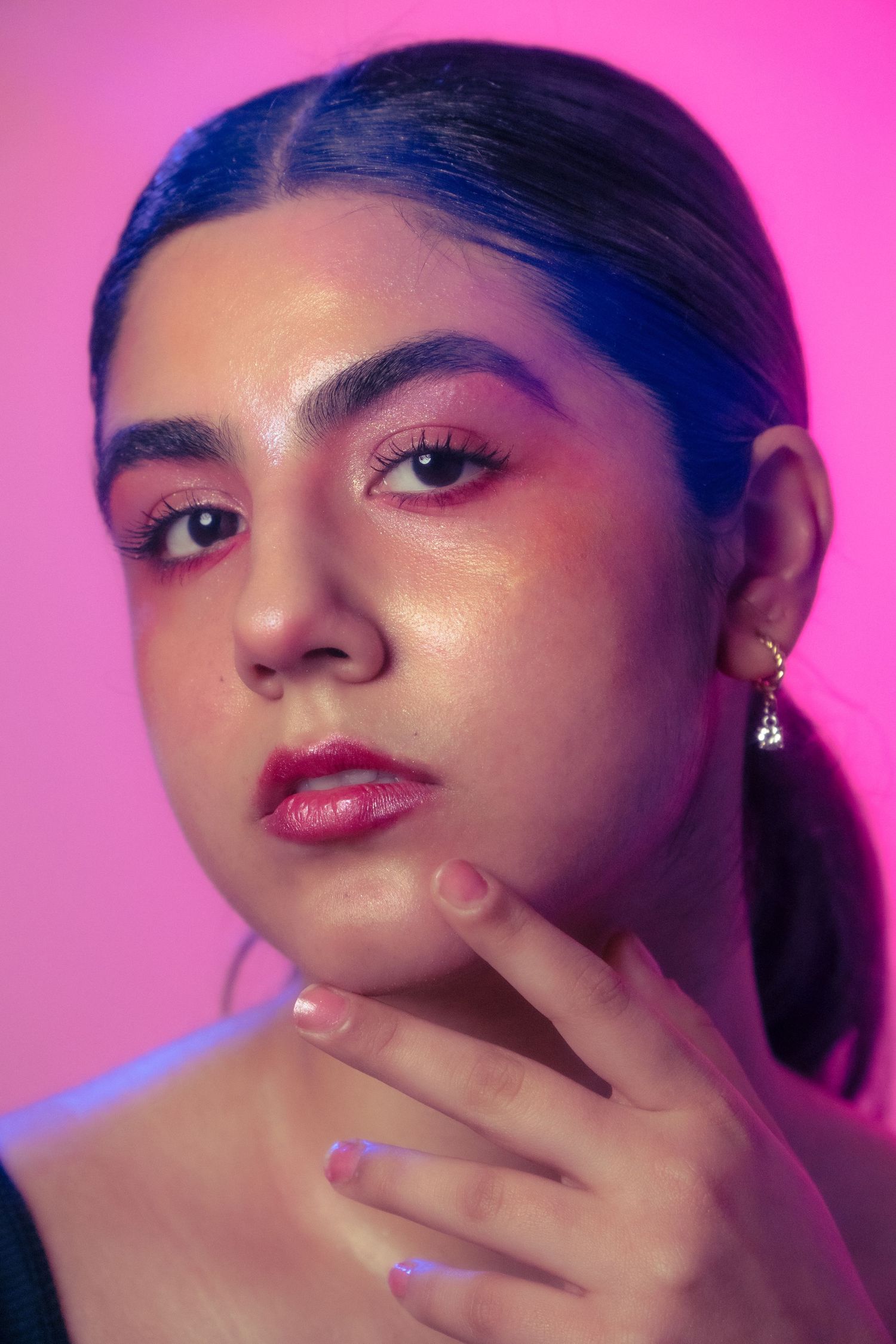 Beauty portrait of young woman with bold pink eyeshadow and red lips gently touching her chin in neon pink light.