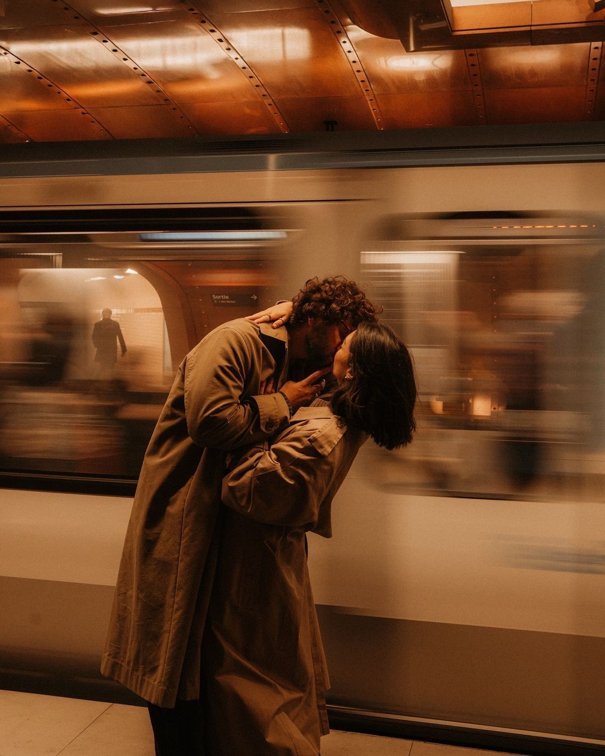 Couple sharing a romantic kiss on a subway platform as a blurred train passes behind them.