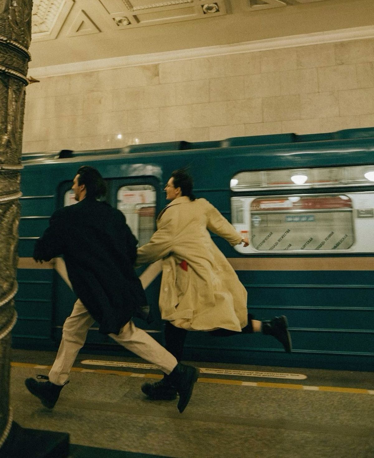 Two people running on a subway platform past a teal train in an ornate underground station.