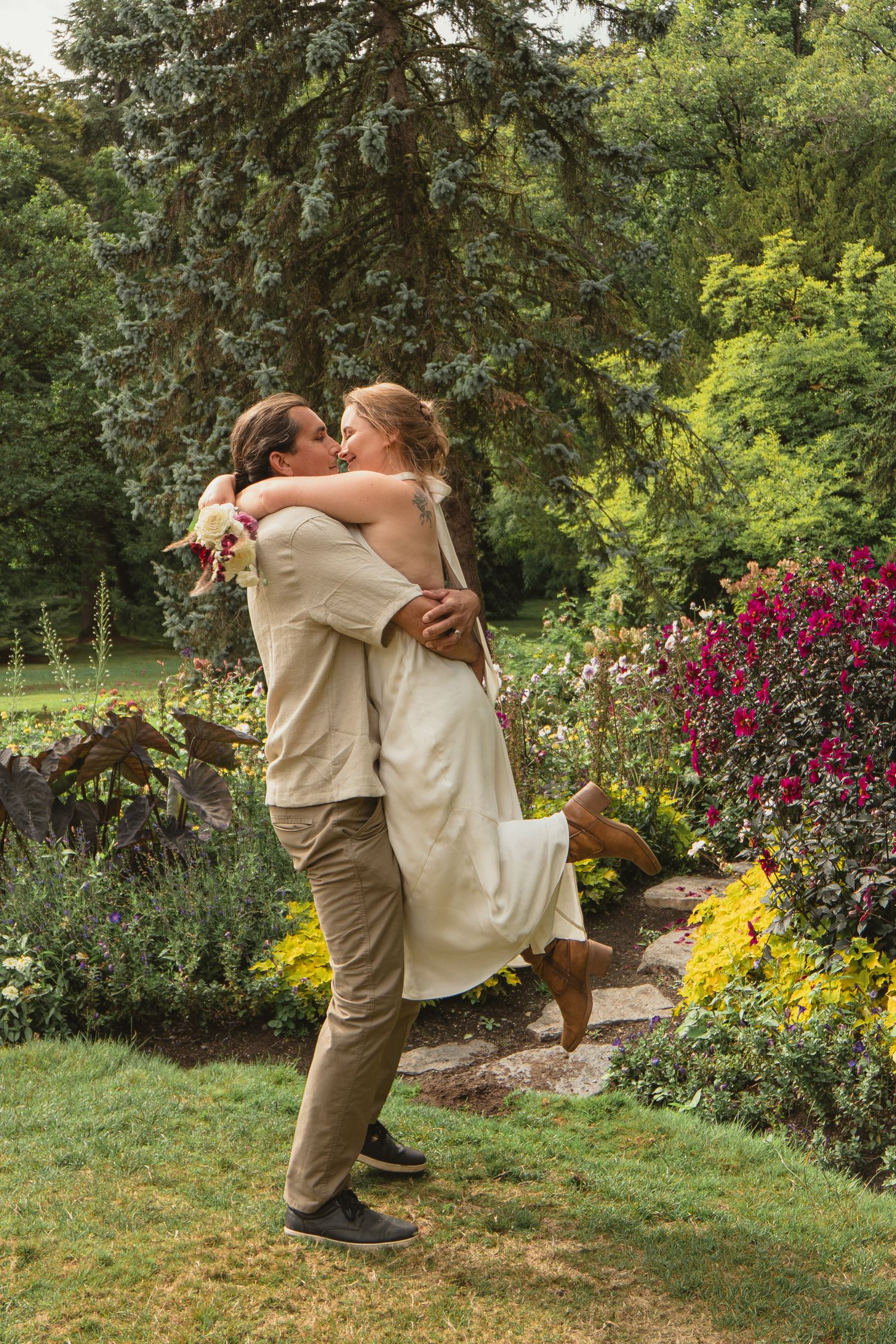 Groom lifting bride in white dress while kissing in a colorful garden landscape.