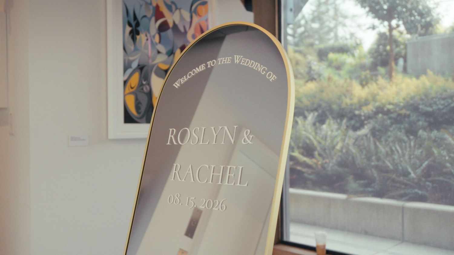 Gold oval mirror displaying wedding welcome sign for Roslyn and Rachel, set against greenery backdrop.