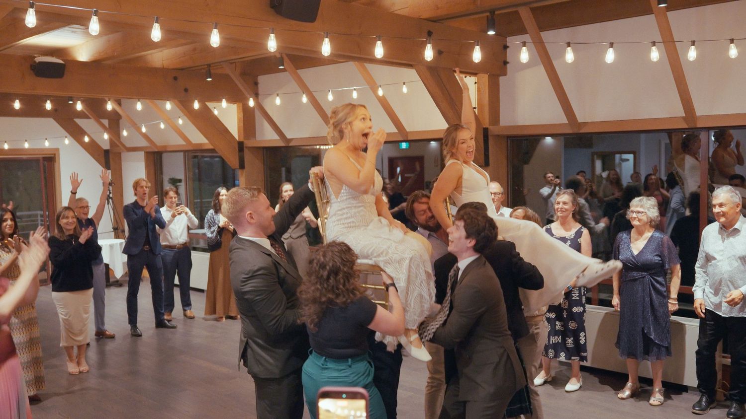 Bride being lifted by guests during a joyful wedding reception dance in an elegant wooden venue.
