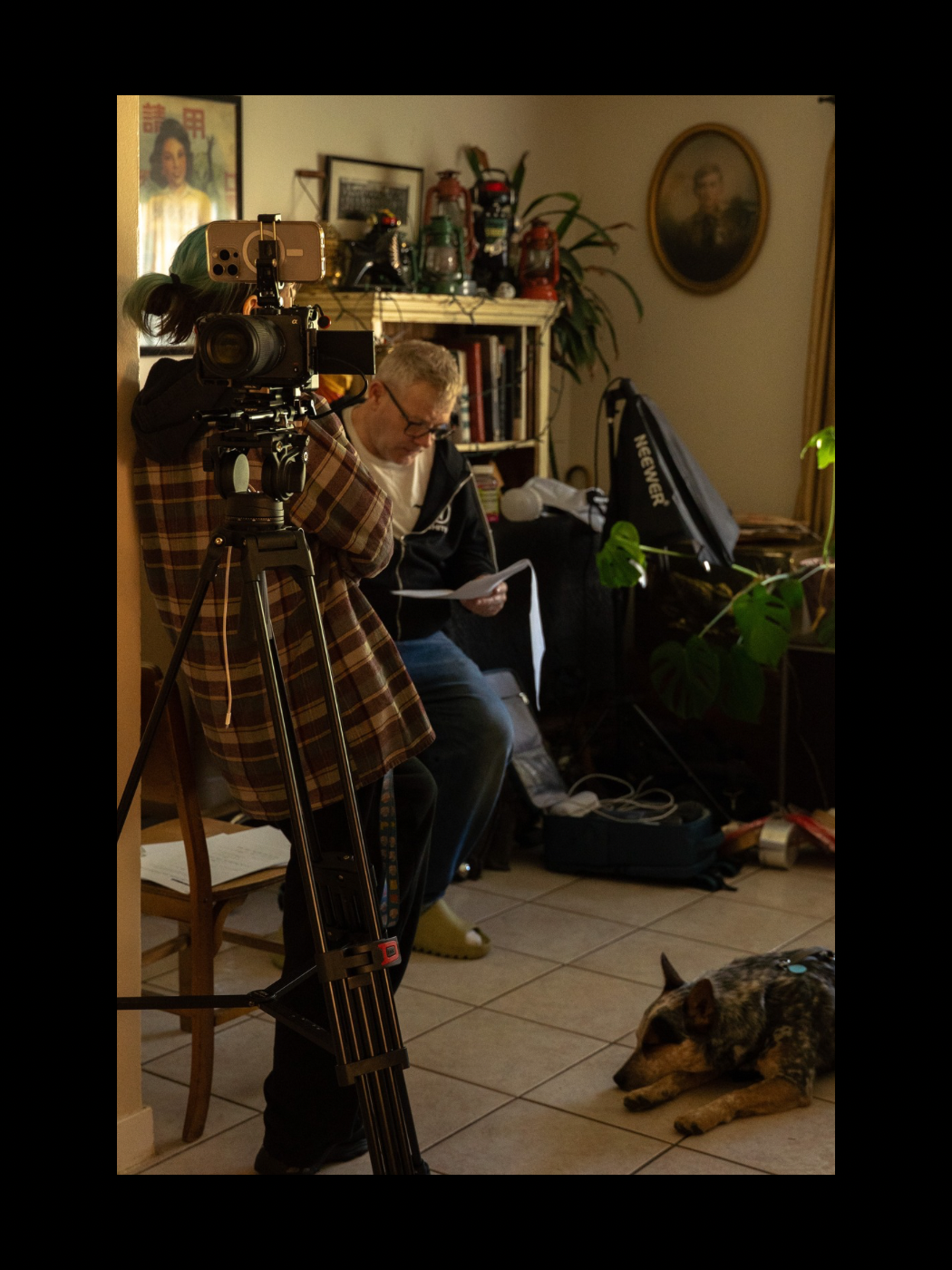 Filmmaker reviews notes beside a professional camera on tripod in a home living room with a dog resting on the floor.