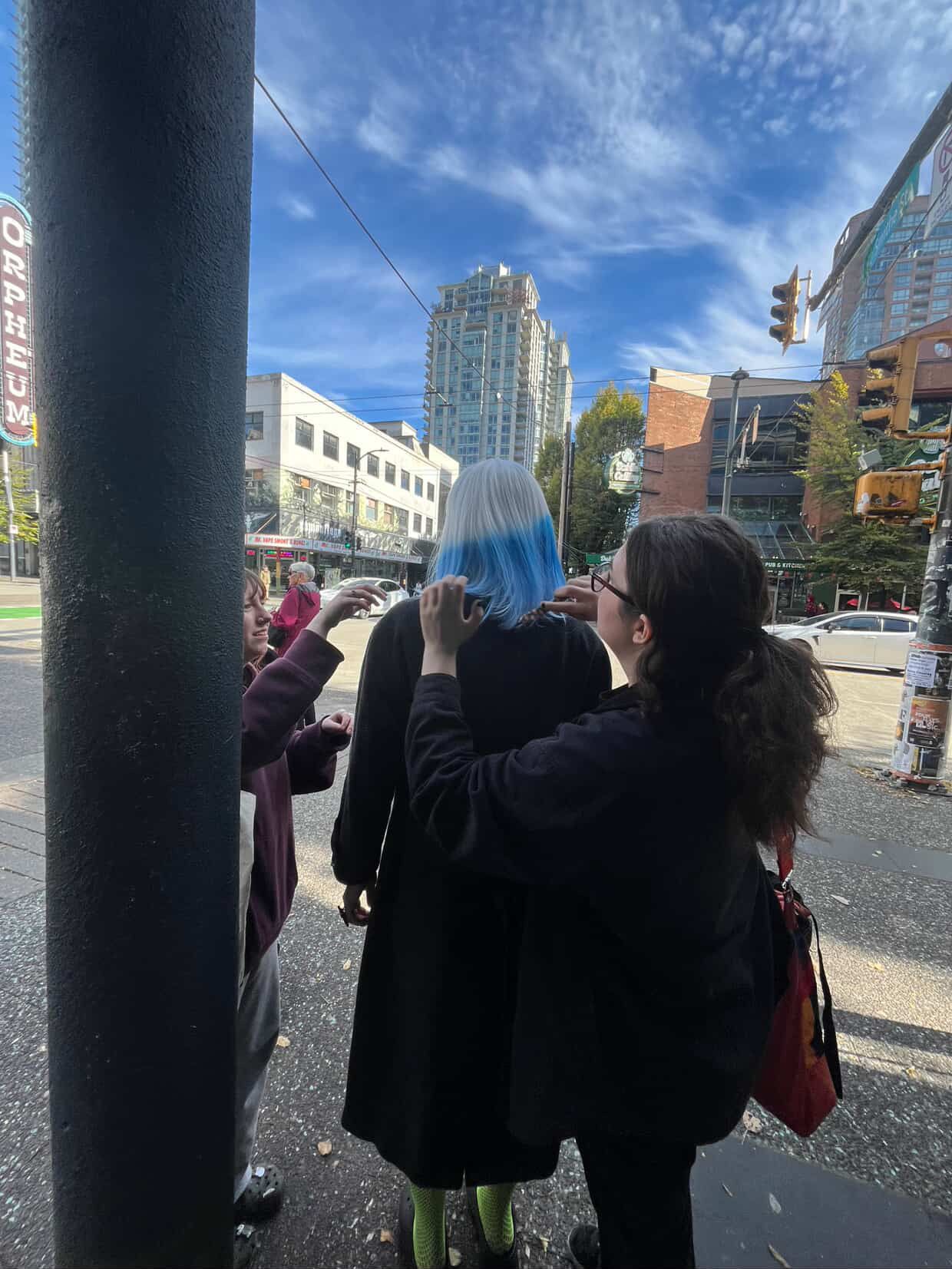 People helping someone put on a blue and white wig on a sunny urban street corner.