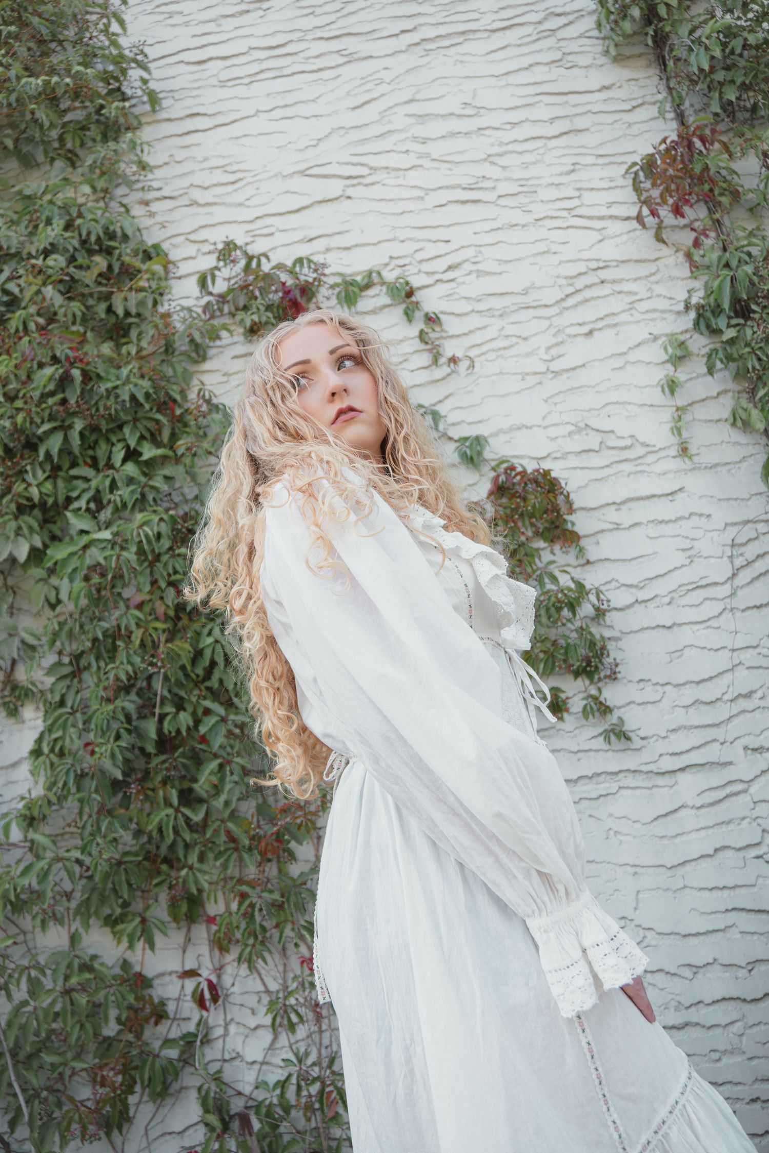 Young woman with long blonde curly hair wearing a white Victorian dress posed against a white textured wall with ivy.