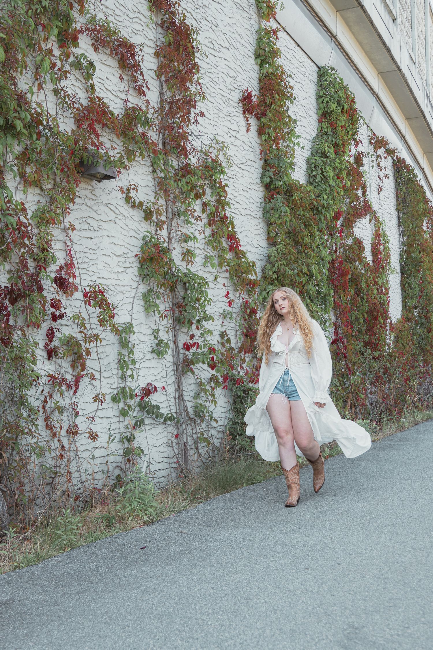 Young blonde woman in white lace duster, denim shorts, and cowboy boots posing by ivy-covered brick wall.