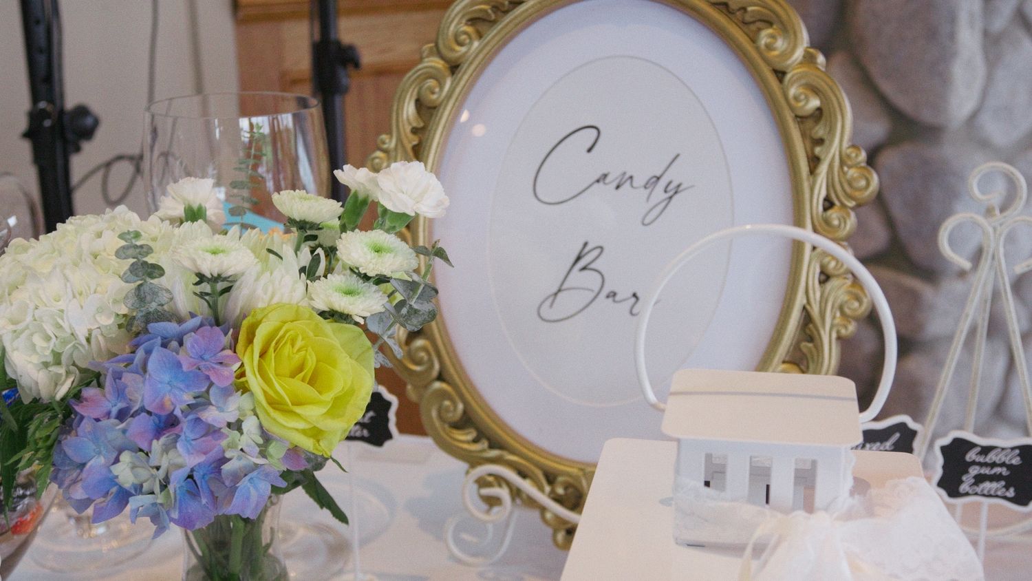 Elegant candy bar display with gold ornate frame sign, blue hydrangeas, yellow roses, and white floral arrangement.