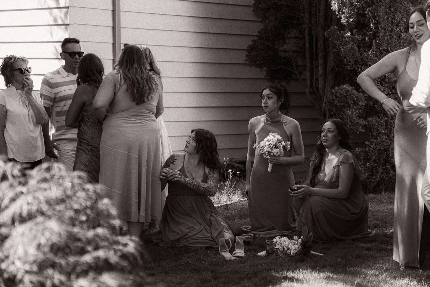 Black and white photo of bridesmaids in long dresses gathered outside a house, some sitting on grass, holding a bouquet.