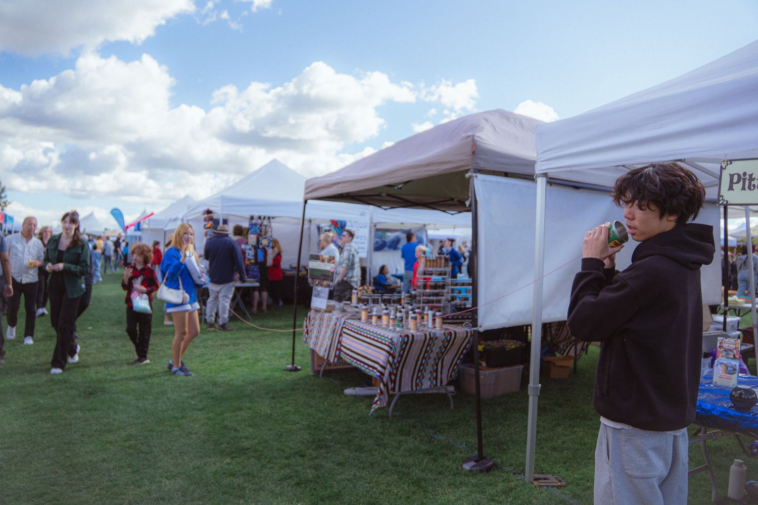 People browse vendor booths under white canopy tents at an outdoor community festival.