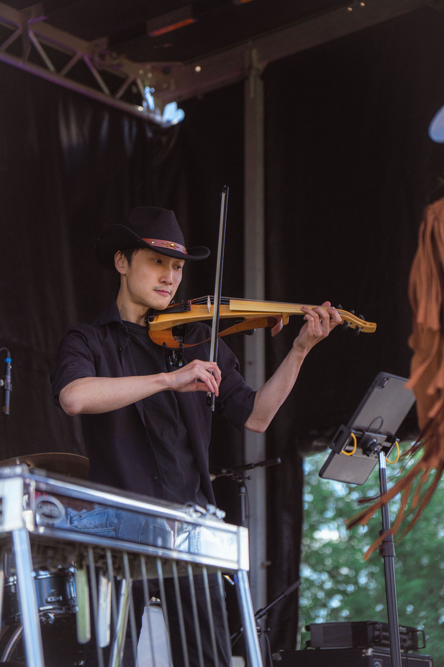 Male musician in black cowboy hat plays electric violin on darkened outdoor festival stage.