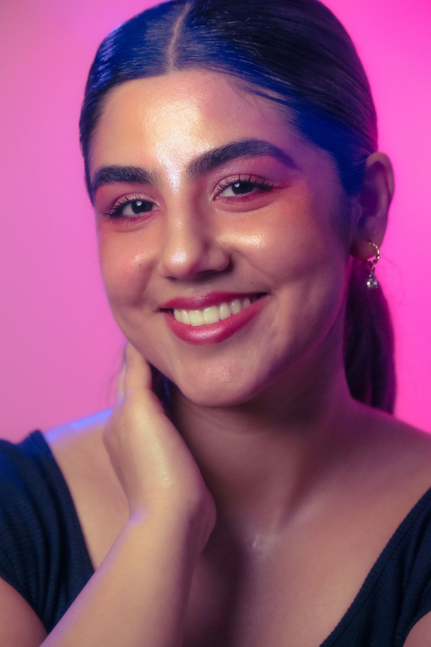 Smiling young woman with pink eye makeup and red lips resting hand on neck against bright pink neon background.