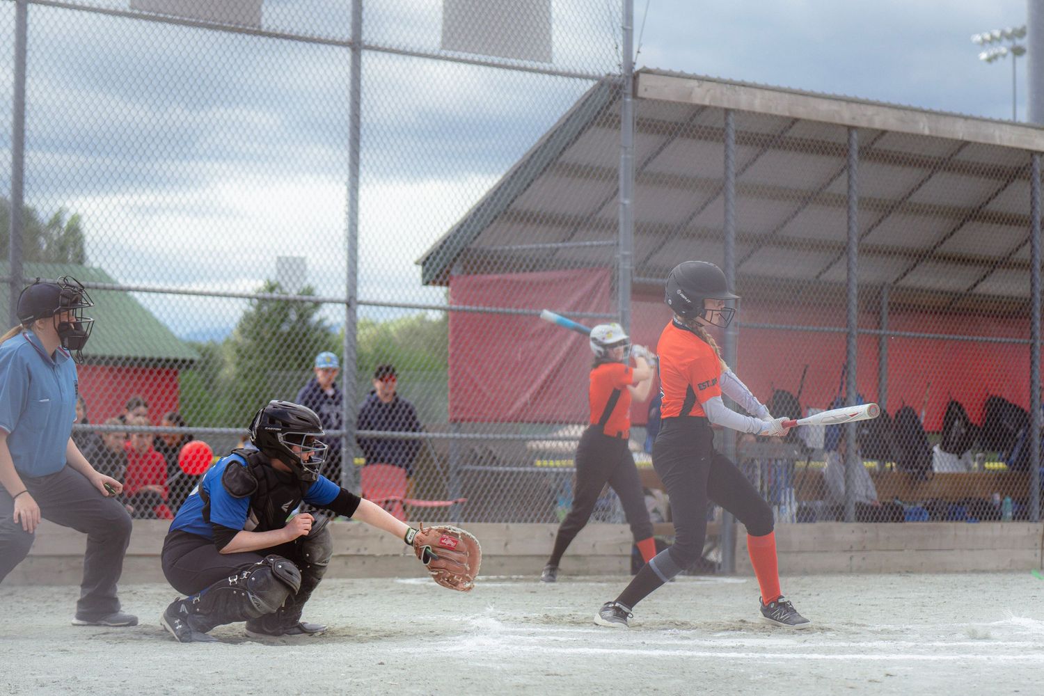 Softball batter in orange uniform hits a pitch as the catcher and umpire watch during a game.
